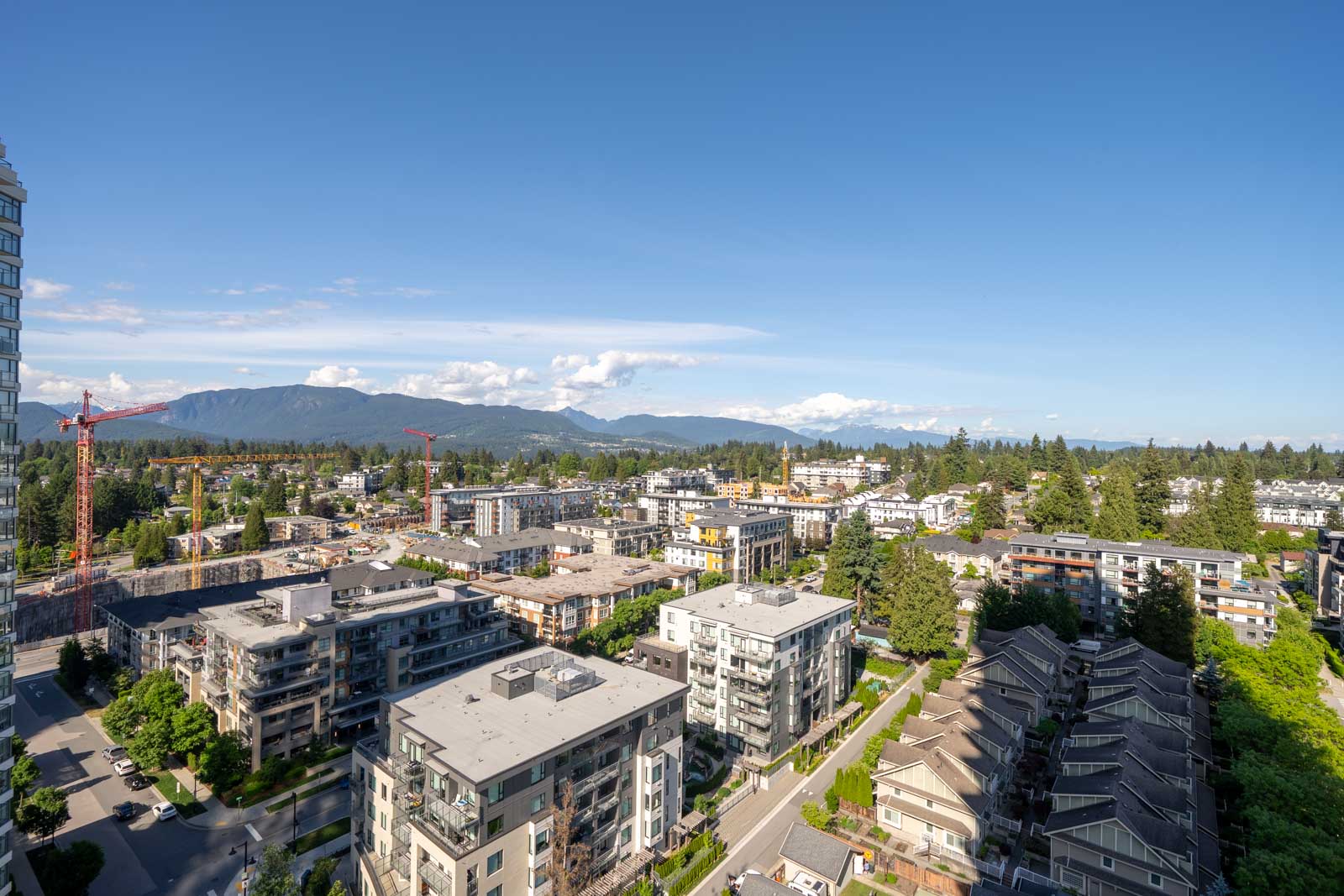 Aerial view of a modern suburban neighborhood with apartment buildings, townhouses, construction cranes, and mountains in the background under a clear blue sky.