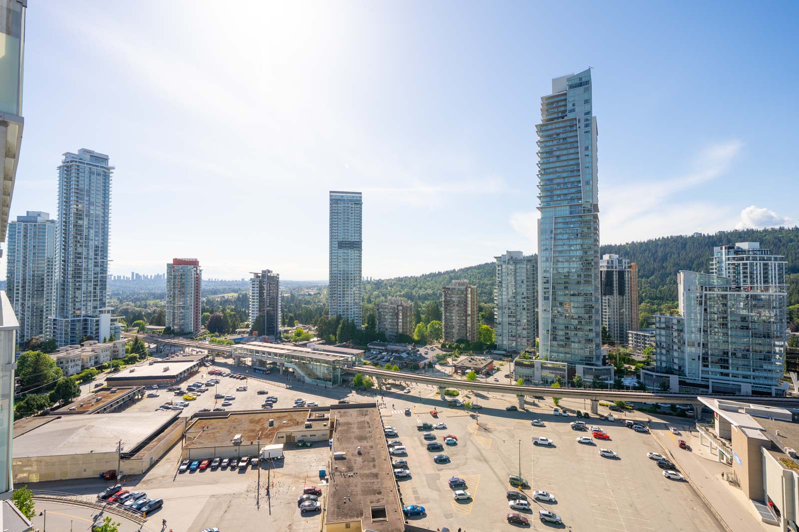 High-rise buildings and a busy road surround a large parking lot under a clear, sunny sky with distant green hills in the background.