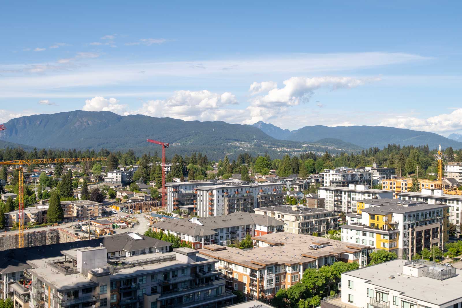 Aerial view of a suburban neighborhood with mid-rise buildings, construction cranes, and forested mountains in the background under a blue sky.