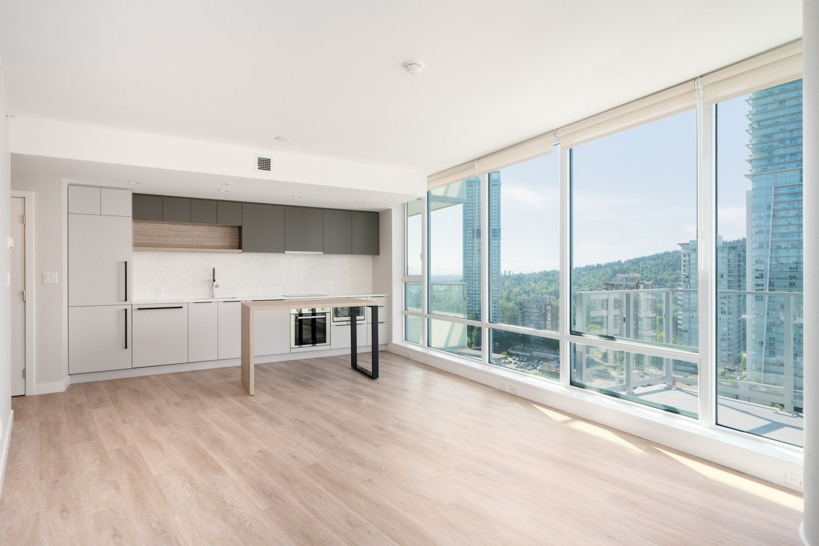 Bright modern apartment with light wood flooring, floor-to-ceiling windows, and an open kitchen, offering a view of city buildings and greenery outside.