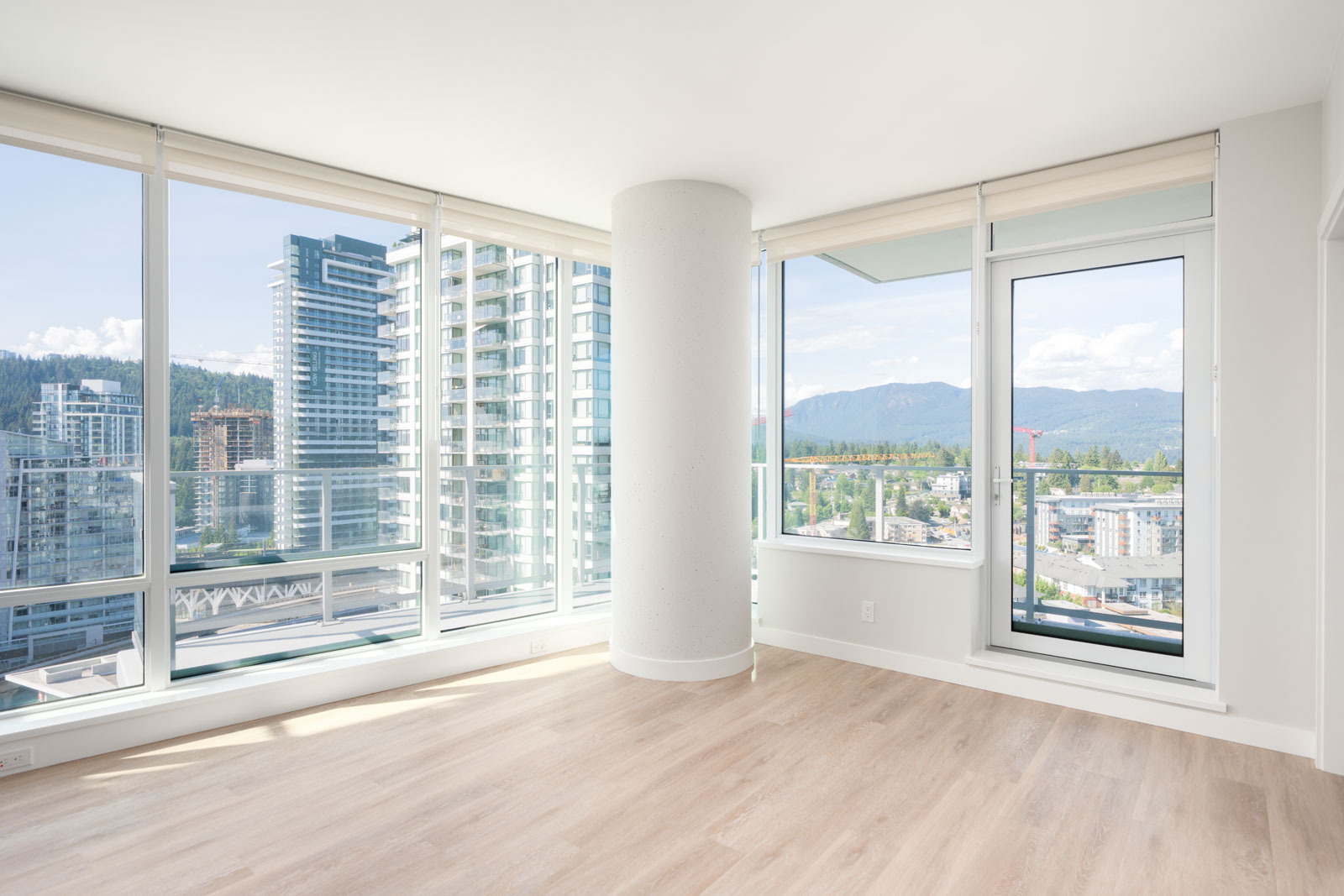 Bright, modern corner apartment with large windows, light wood flooring, a round pillar, and a view of city buildings and mountains.