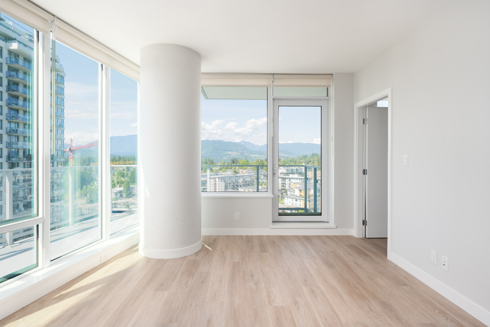 Bright empty room with large windows, a glass door to a balcony, a round pillar, and light wood flooring. Cityscape and mountains visible outside.