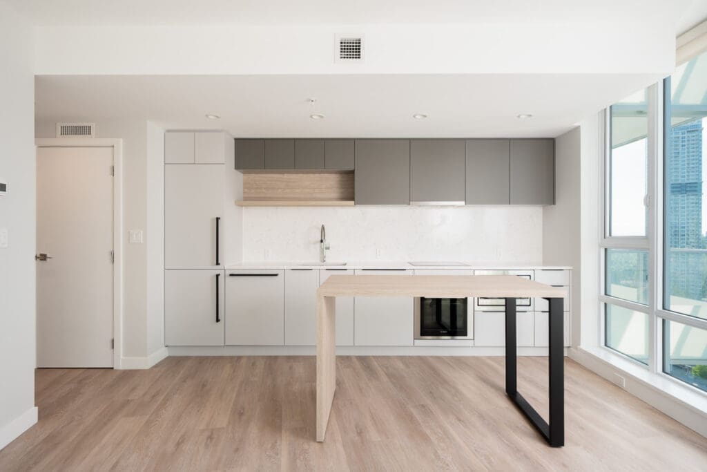 Minimalist modern kitchen with light wood flooring, gray cabinets, built-in appliances, and a light wood island with a black metal support, next to large floor-to-ceiling windows.