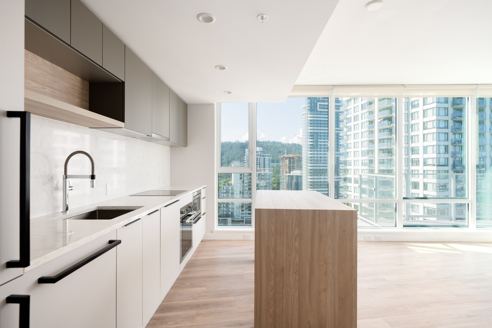 Modern kitchen with light wood island, built-in appliances, and large windows offering a city view with high-rise buildings and natural light.