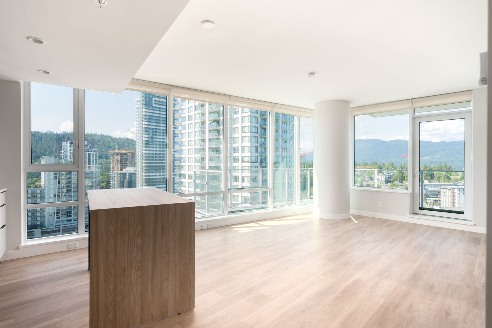 A modern, unfurnished apartment with large floor-to-ceiling windows, light wood flooring, and views of surrounding high-rise buildings and mountains.