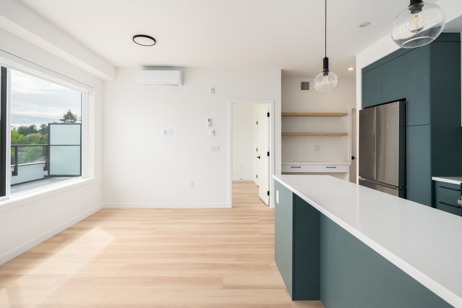 Modern, unfurnished kitchen and dining area with light wood floors, white walls, green cabinets, pendant lights, and large window providing natural light.