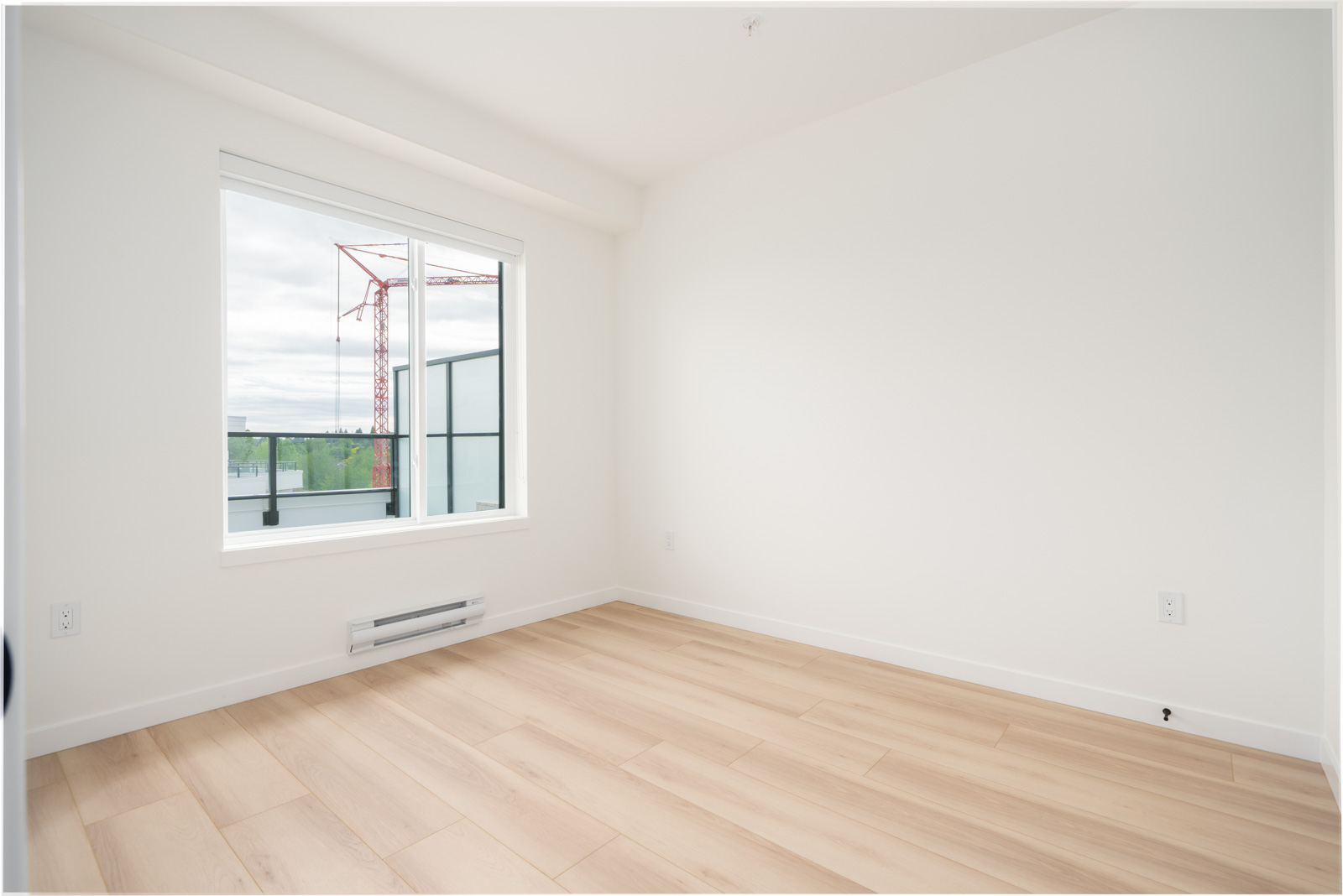 Empty room with light wood flooring, white walls, a window with a view of a construction crane, and a baseboard heater below the window.