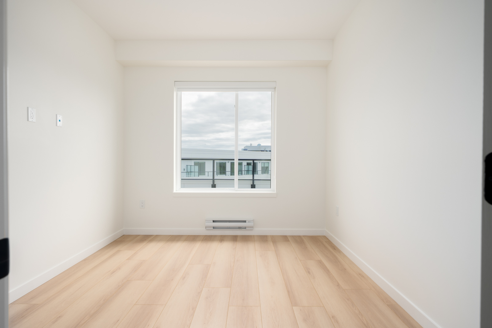 Empty, unfurnished room with light wood flooring, white walls, and a window showing an exterior building and cloudy sky.