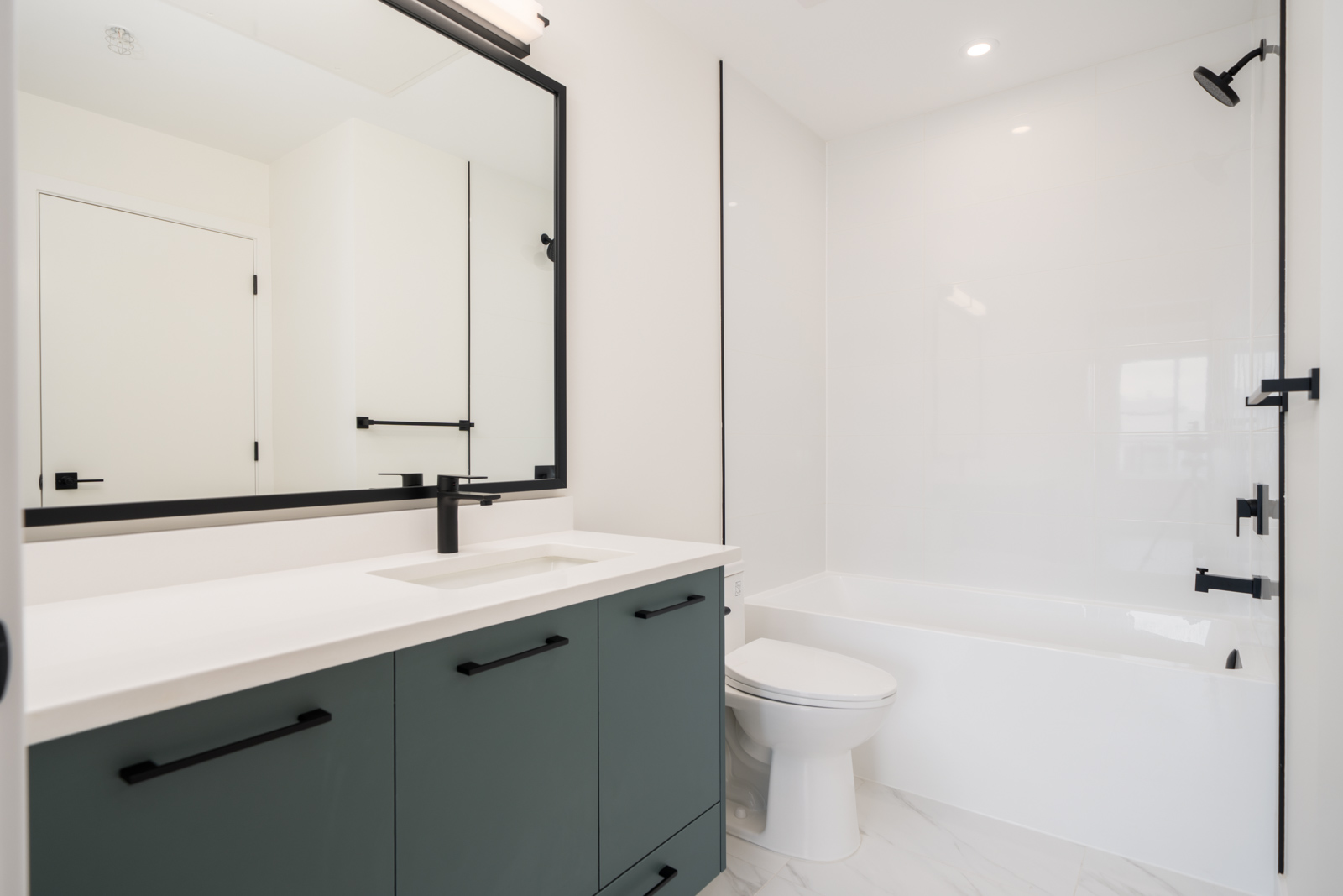 Modern bathroom with a large mirror, green vanity, white countertop, black fixtures, toilet, and a bathtub with a shower.