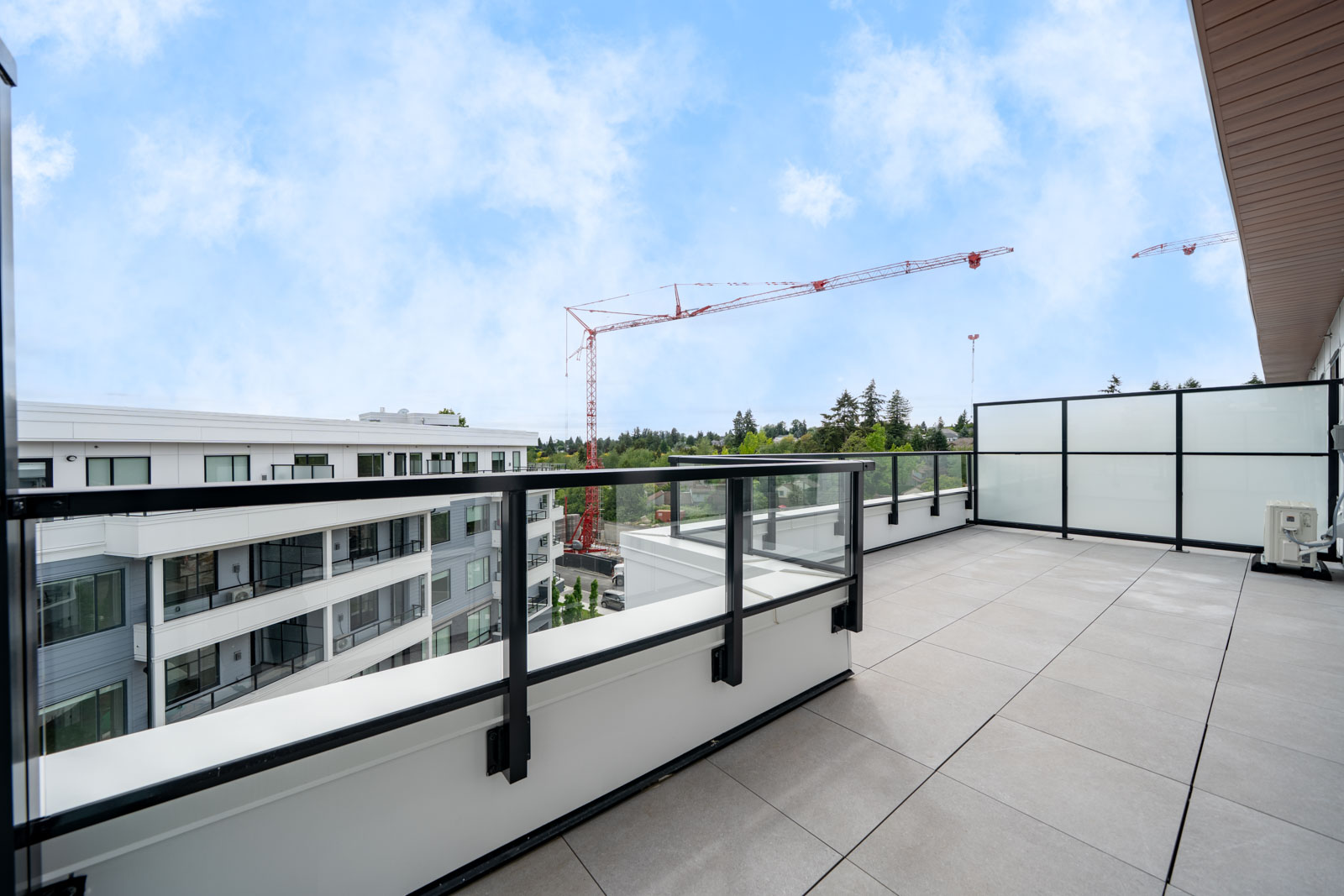 Large balcony with glass railings overlooks a modern apartment complex; construction cranes and trees are visible in the background under a partly cloudy sky.