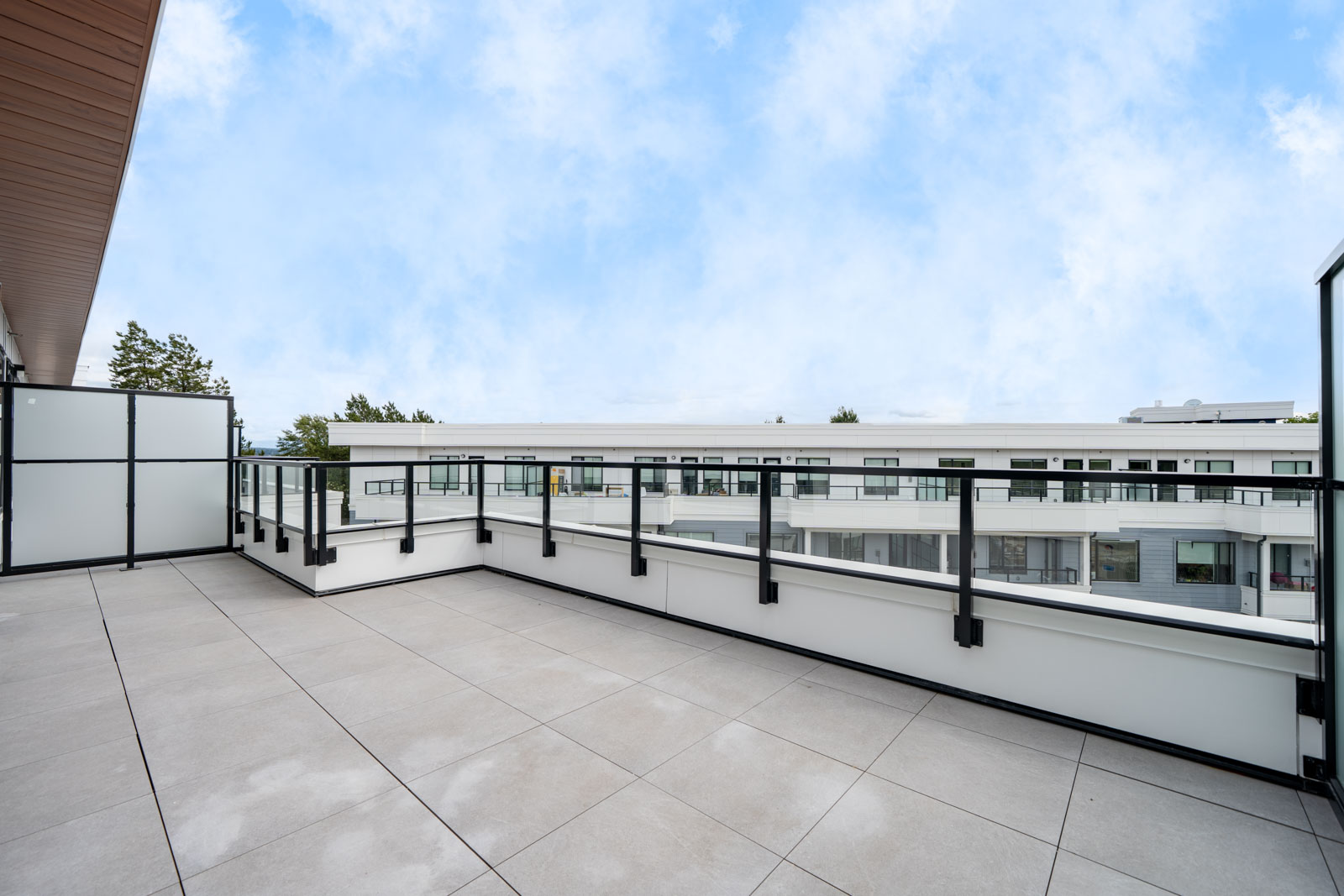 Spacious rooftop terrace with tiled flooring, glass railing, and view of modern apartment buildings under a partly cloudy sky.