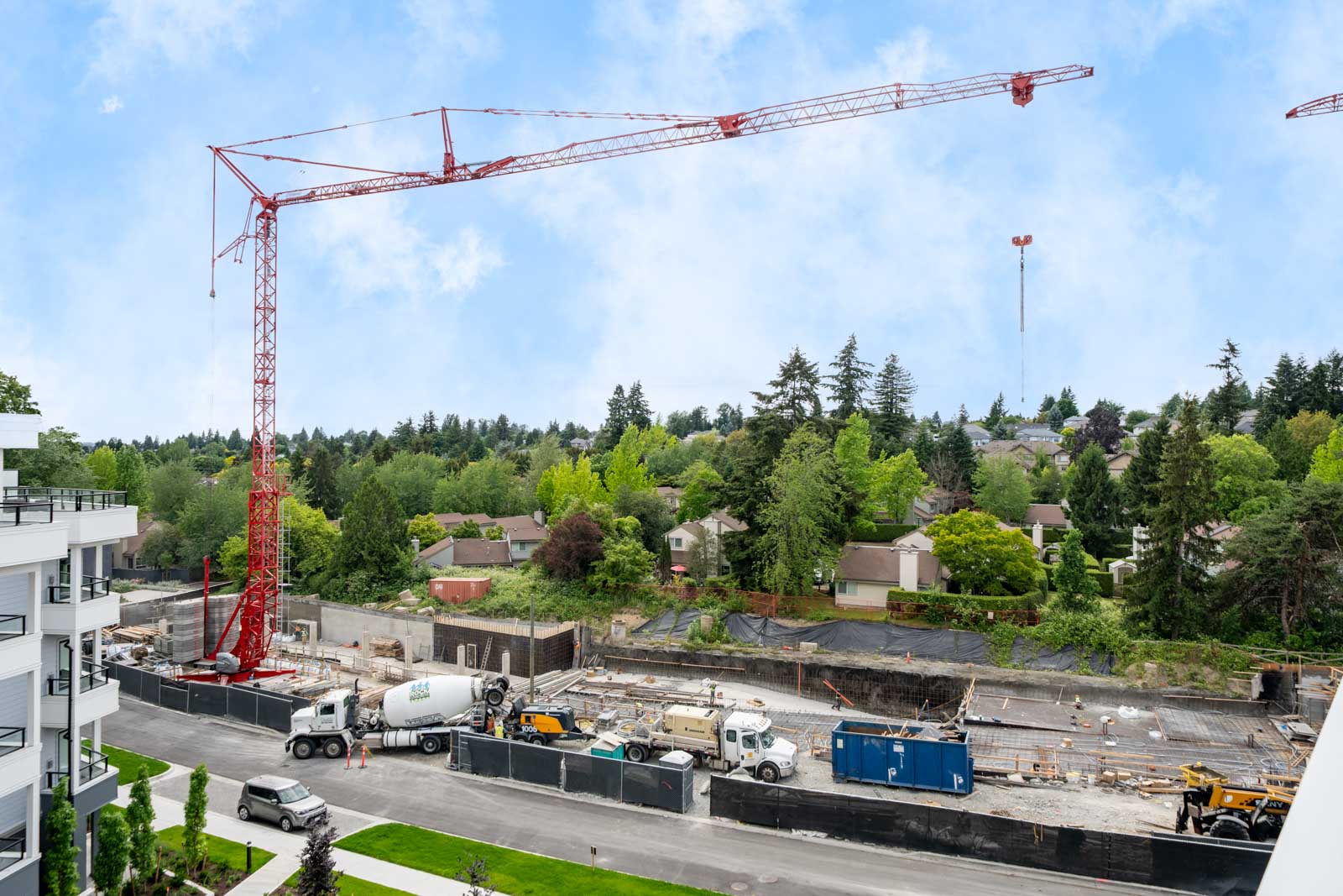 A construction site with a red crane, cement trucks, and building materials, set near a residential area with trees in the background.