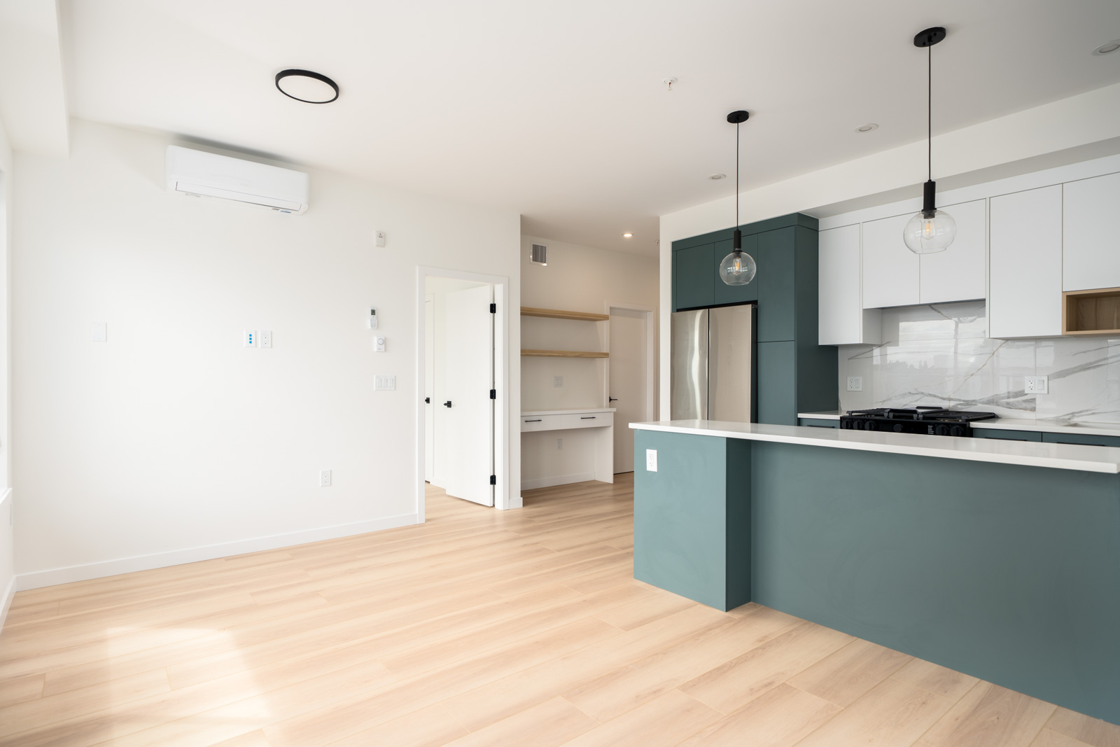 Modern kitchen with teal island, white cabinets, stainless steel appliances, light wood floors, and a built-in desk area in the adjacent room. The space is bright and uncluttered.