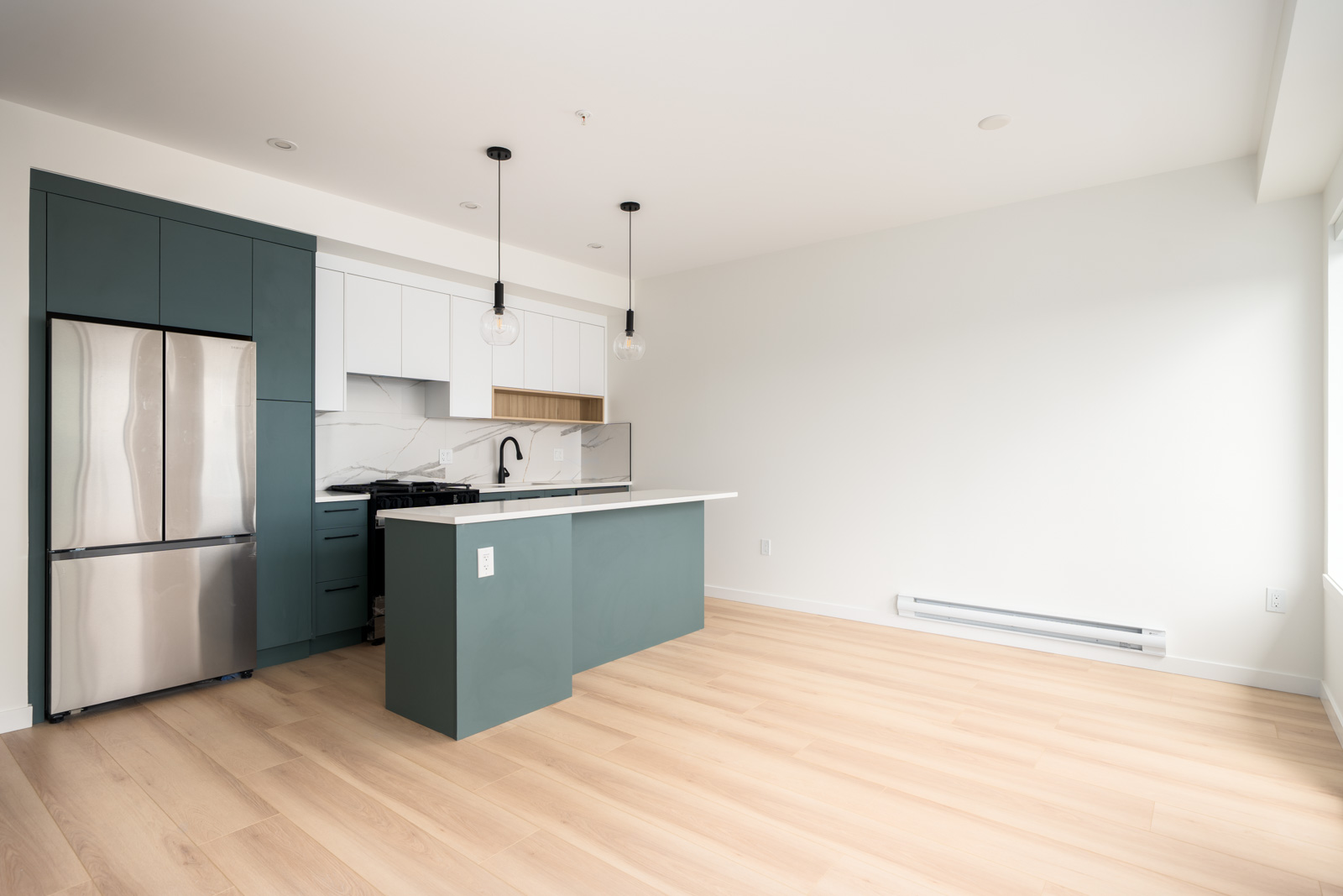 Modern, unfurnished kitchen with green cabinets, stainless steel appliances, a central island, pendant lights, and light wood flooring in a bright, empty room.