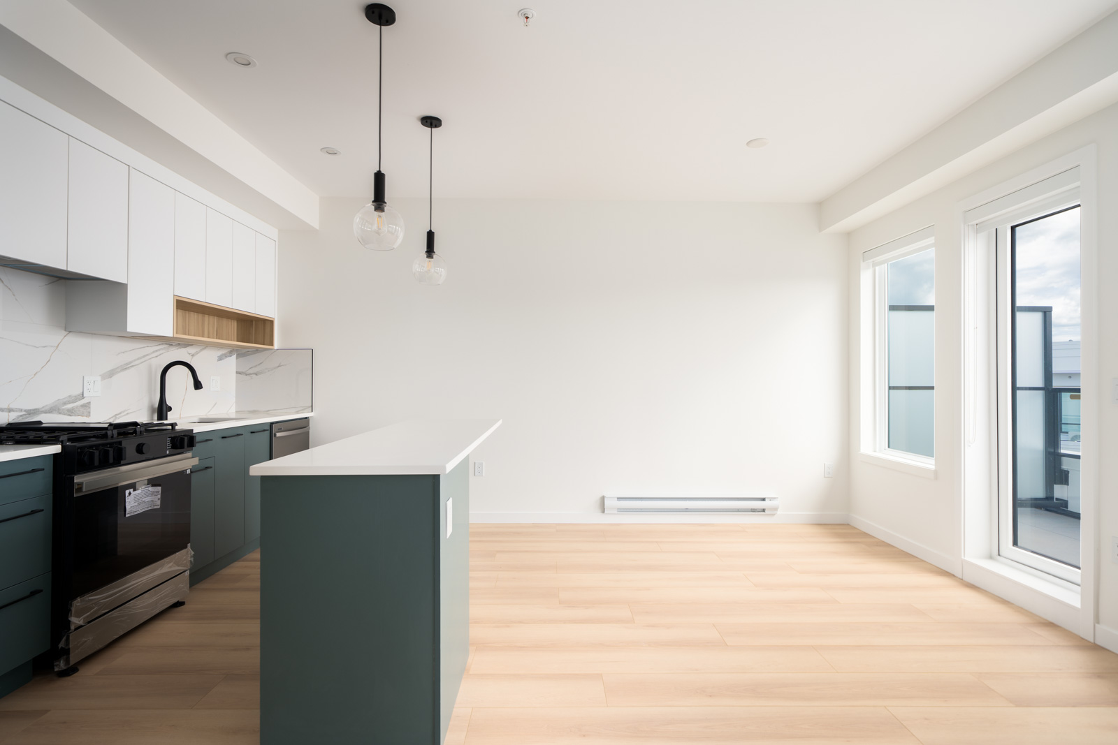 Modern kitchen with white countertops, green lower cabinets, hanging pendant lights, and large windows letting in natural light.