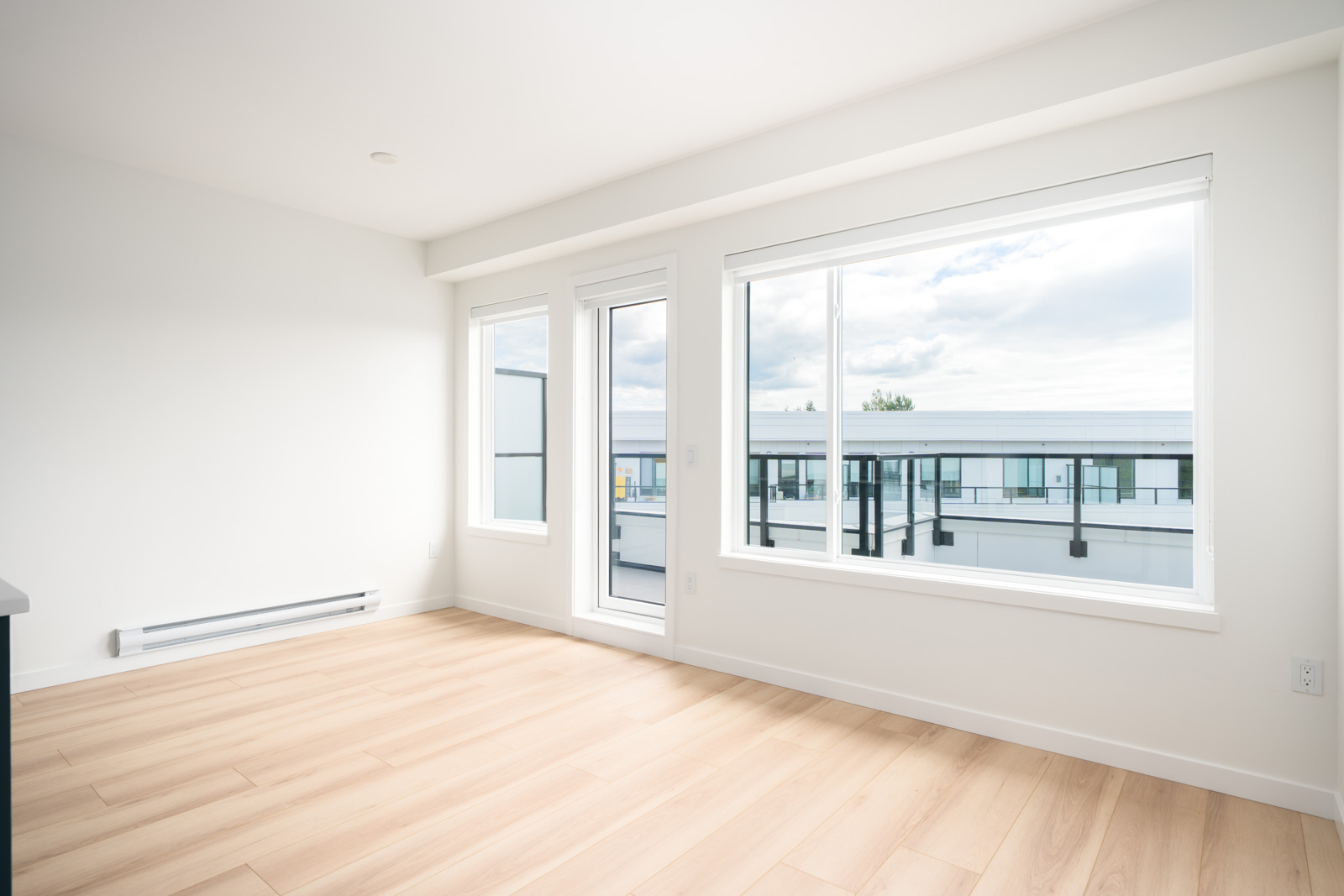 Bright, empty room with light wood flooring, large windows, and a glass door opening to a balcony with a view of modern buildings and a partly cloudy sky.