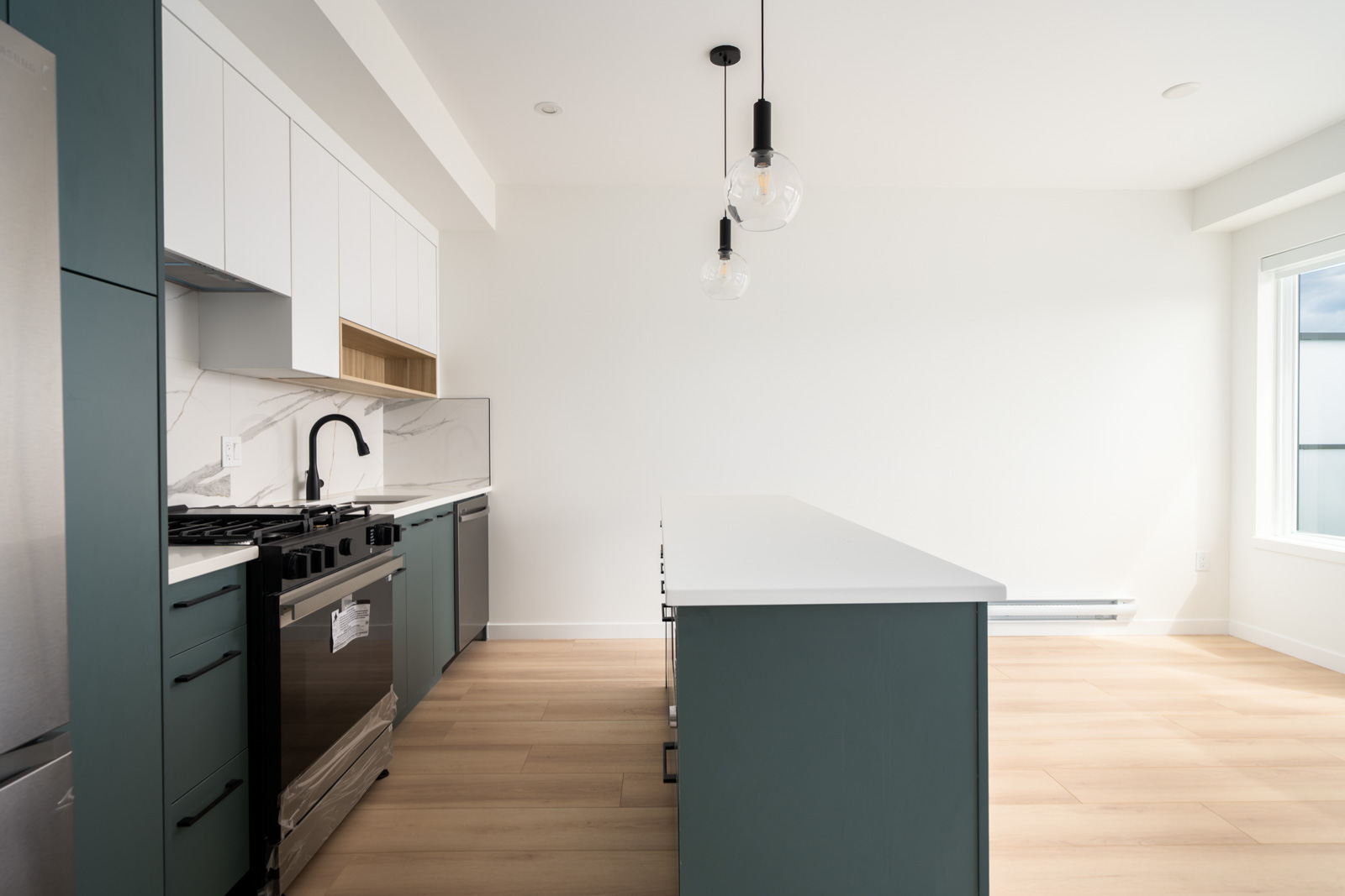 Modern kitchen with teal lower cabinets, white upper cabinets, stainless steel appliances, white countertops, island with pendant lights, and light wood flooring.