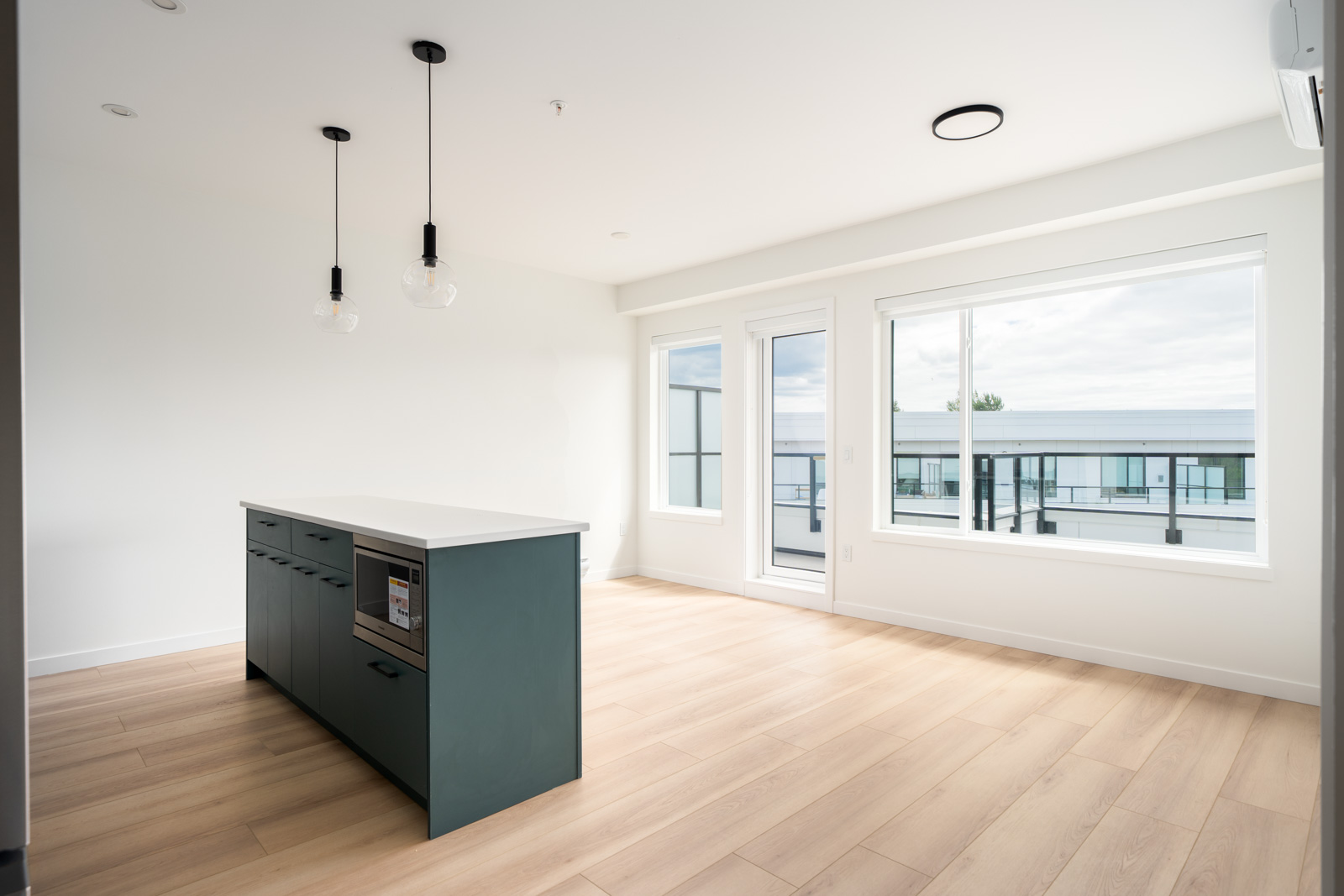 A modern, unfurnished kitchen and living area with light wood flooring, a teal island, large windows, and pendant lights, featuring ample natural light.