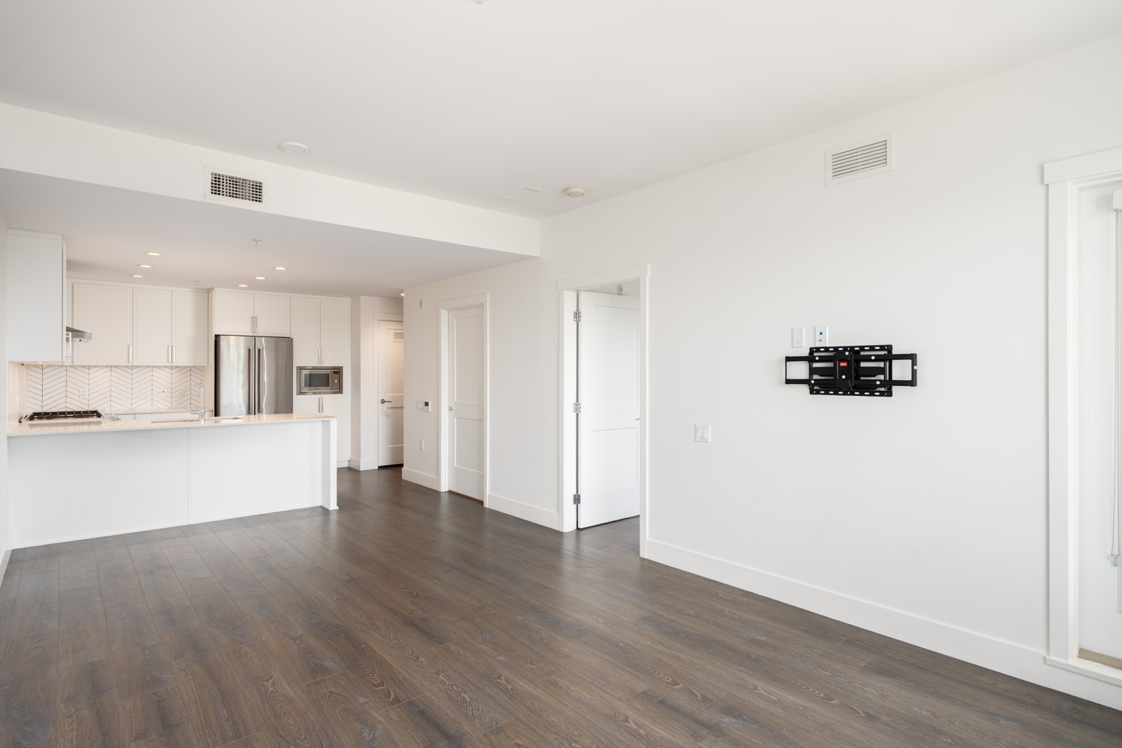 Unfurnished modern apartment with white walls, dark wood flooring, open kitchen area, and a wall-mounted TV bracket.