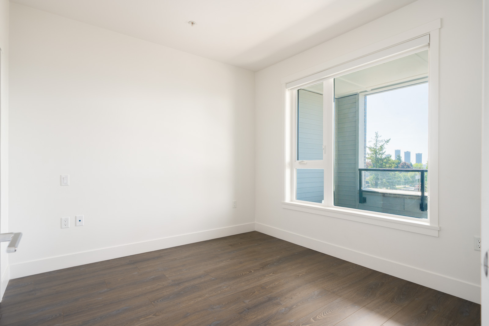 Empty room with white walls, wood flooring, a large window, and a view of buildings and trees outside.