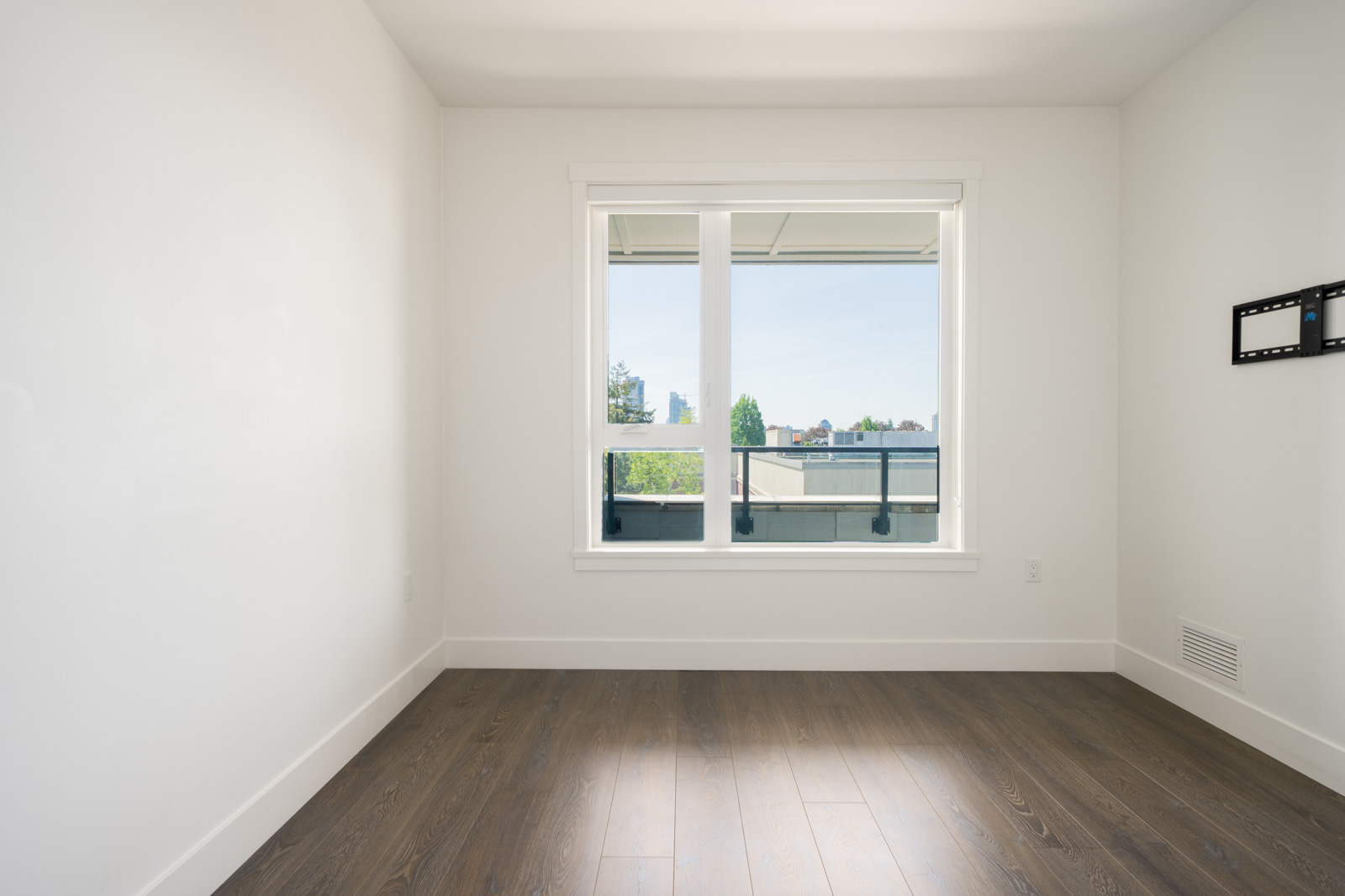 Empty, unfurnished room with white walls, large window, wood floors, and a wall-mounted bracket. Balcony and trees visible through the window.