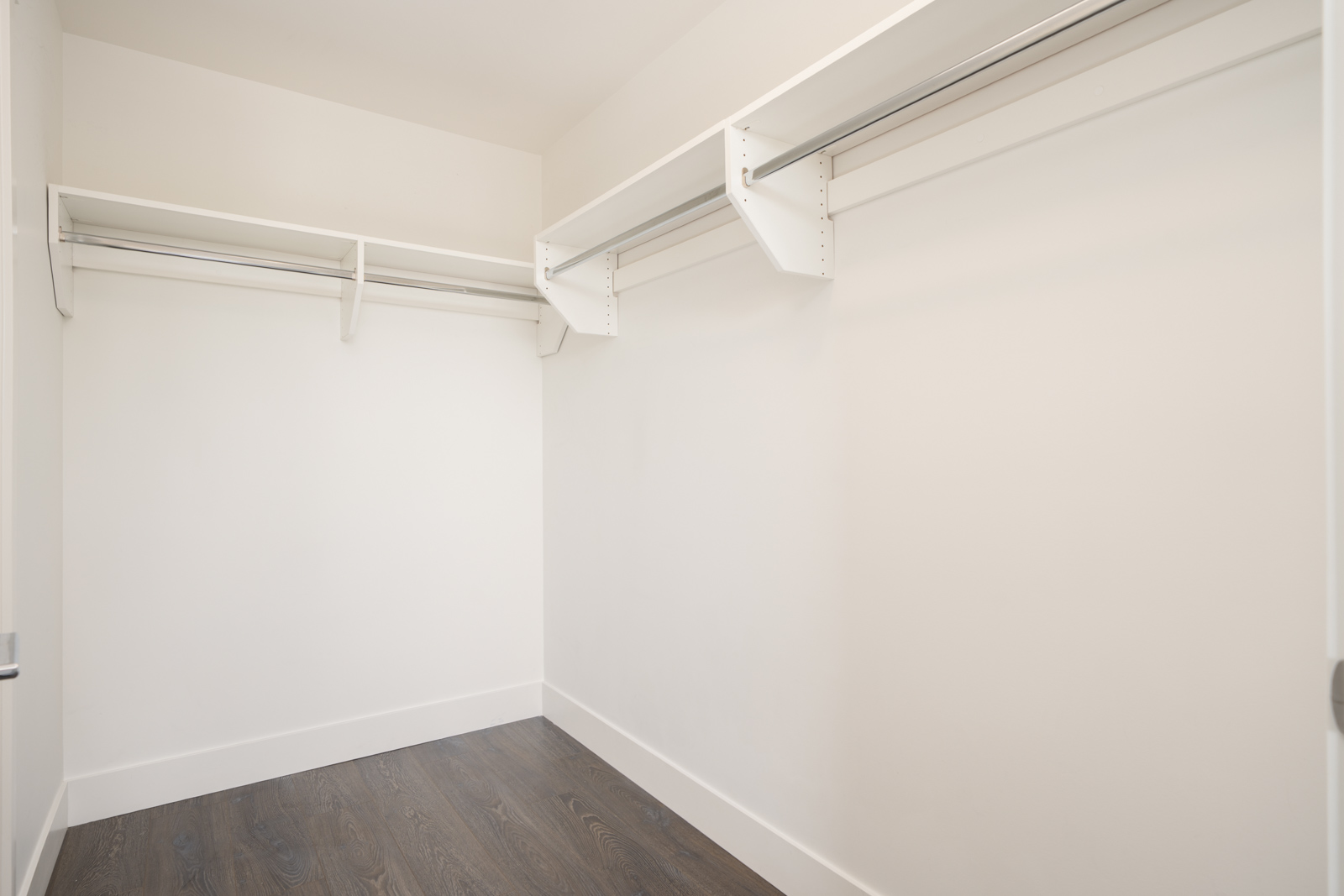 Empty walk-in closet with white walls, shelves, and hanging rods, and dark wood flooring. The space is well-lit and has a clean, minimalist appearance.