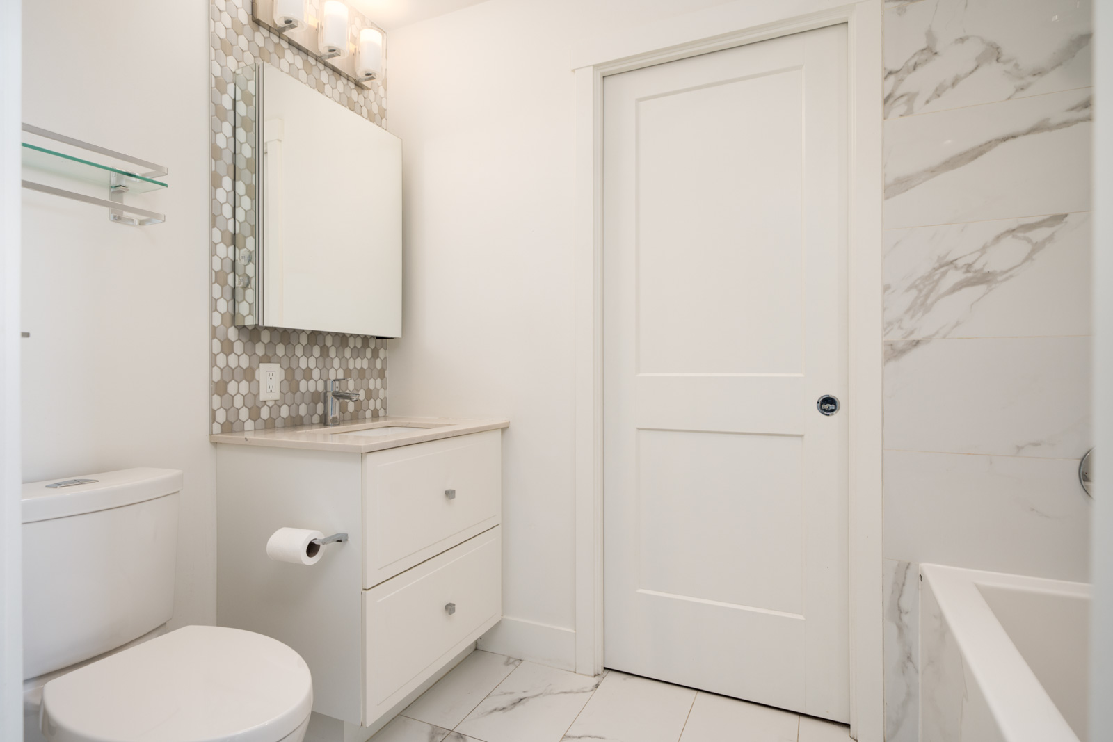 Modern bathroom with a white vanity, toilet, sliding door, and bathtub. Wall tiles have a marble pattern; mosaic tiles are behind the mirror and sink area.