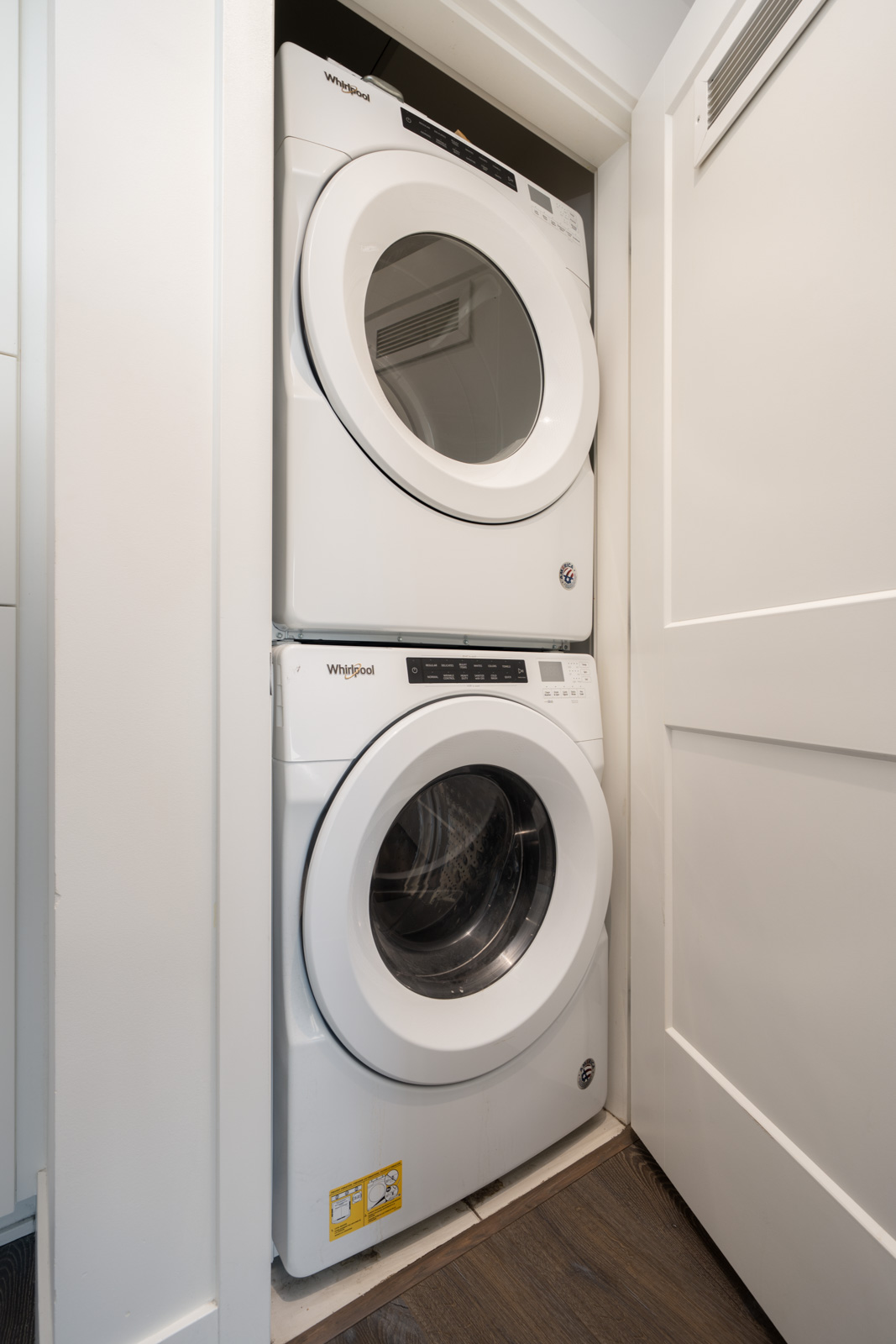 A white Whirlpool washer and dryer are stacked in a narrow laundry closet with a sliding door, positioned on a dark wood floor.