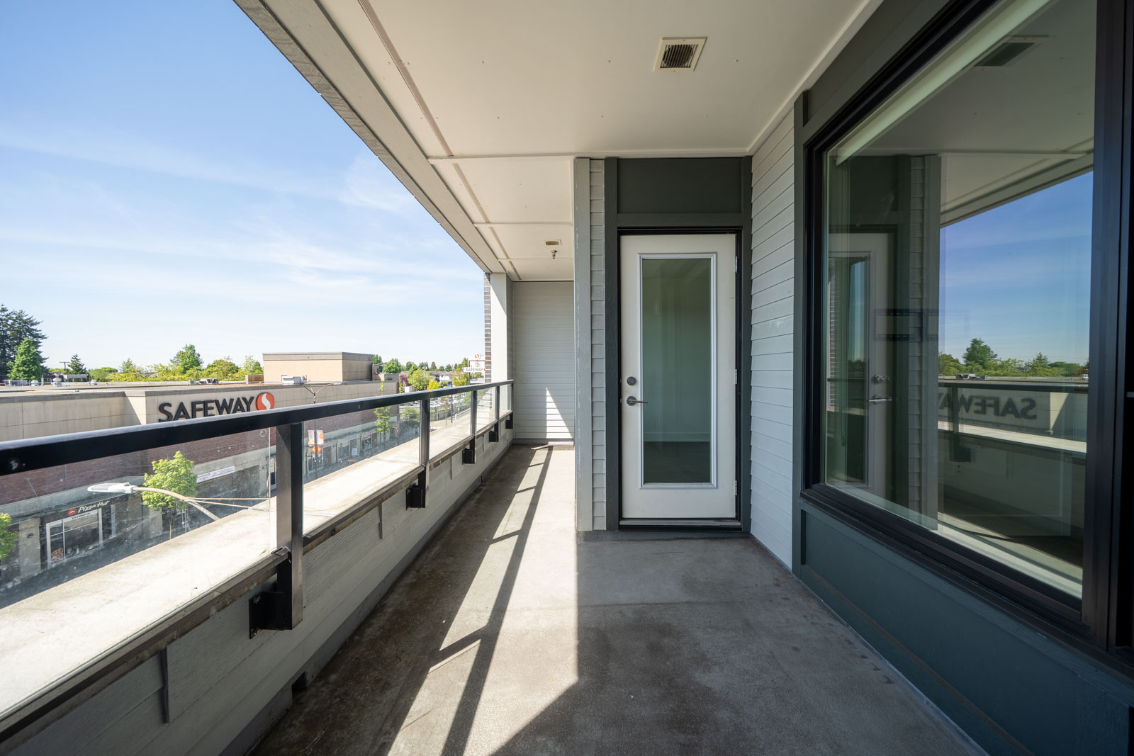 A modern apartment balcony with glass railing, concrete floor, and a glass door, overlooking a Safeway grocery store and nearby buildings.