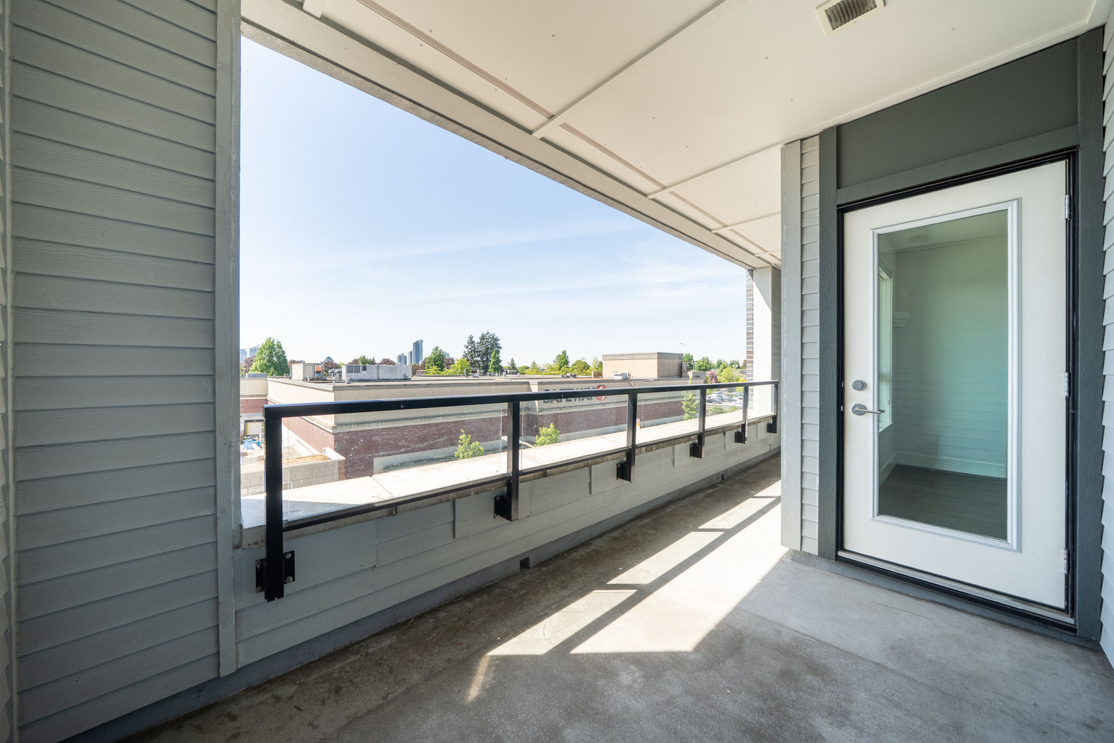 A modern balcony with a glass door, glass railing, and a view of nearby buildings and trees under a clear sky.