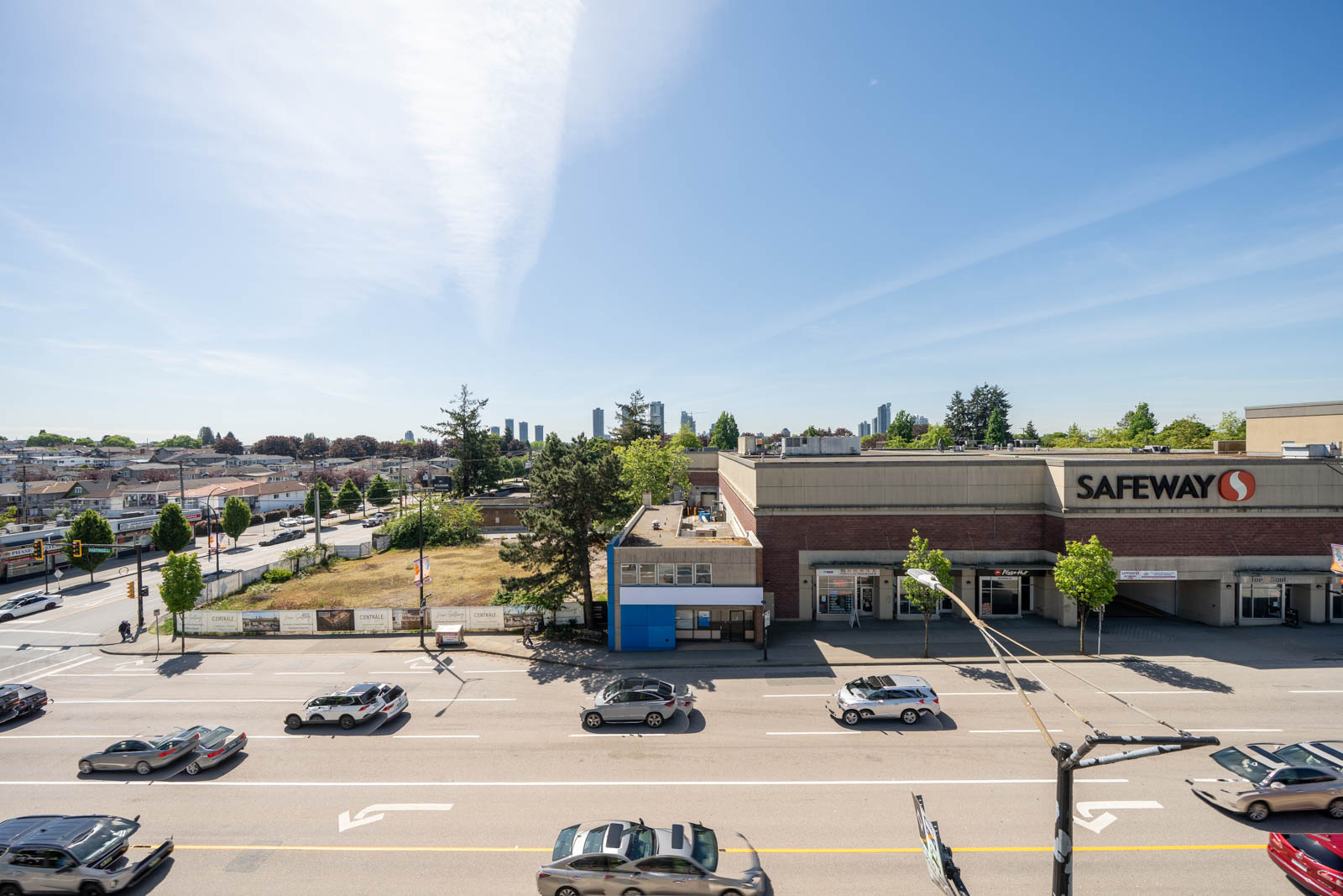 A view of a busy street with cars passing by a Safeway supermarket on the right, under a clear blue sky. Residential buildings and trees are visible in the background.