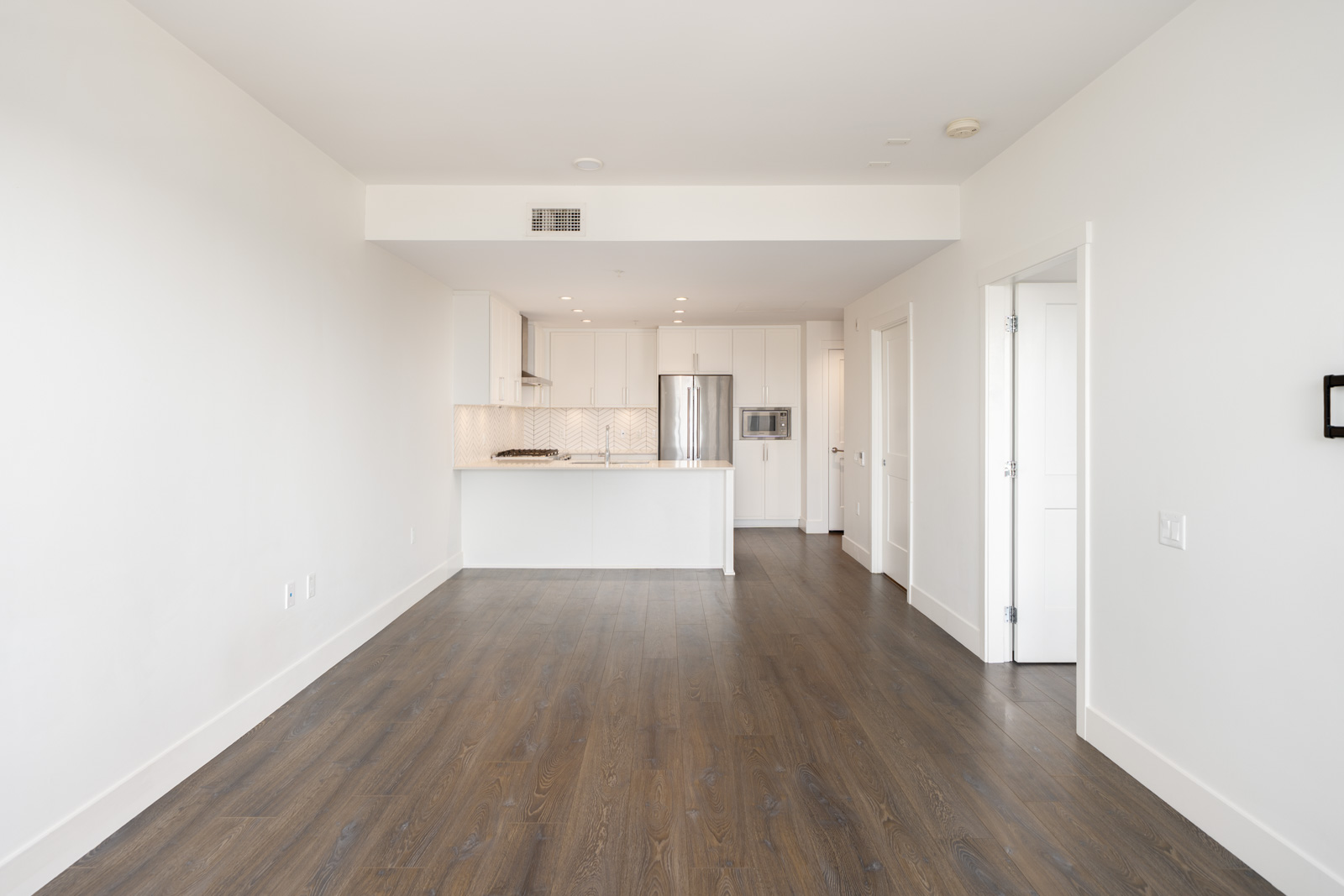 Empty apartment with white walls, dark wood floors, and a modern kitchen featuring white cabinets and stainless steel appliances in the background.