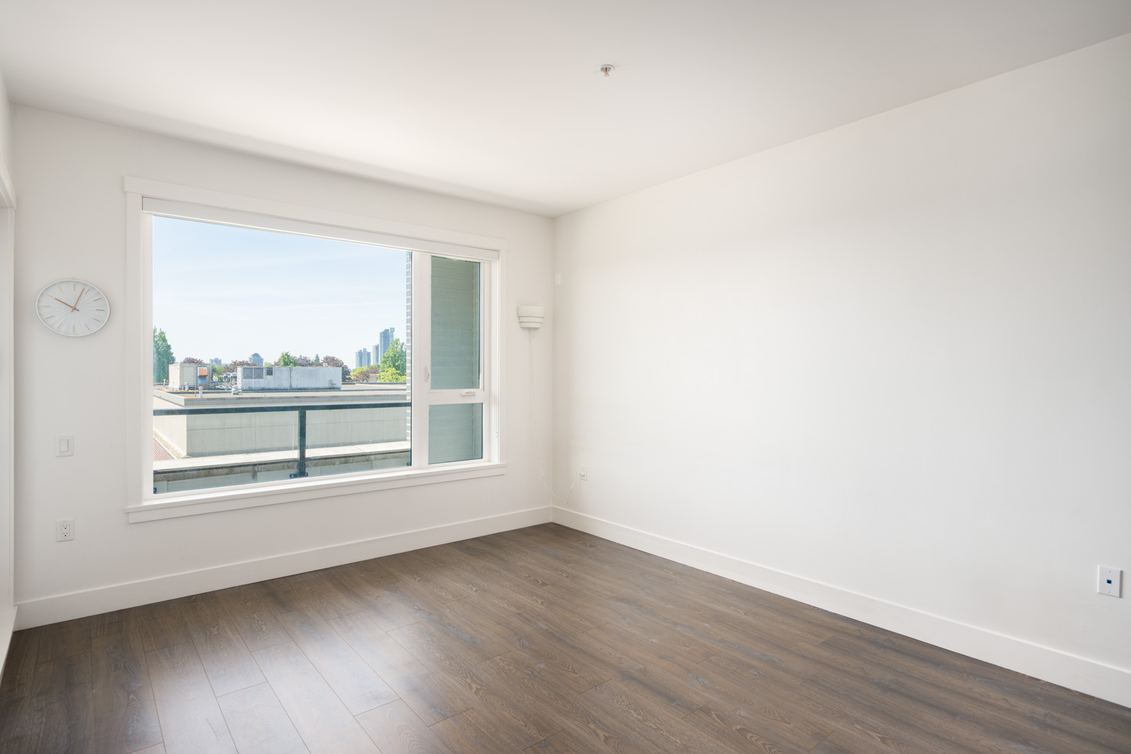 Empty room with white walls, a large window letting in natural light, a wall clock, dark wood flooring, and a view of buildings outside.