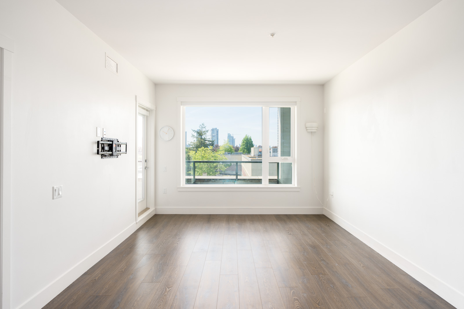 Empty, unfurnished room with white walls, wood flooring, and a large window overlooking a cityscape with trees and buildings in the background.