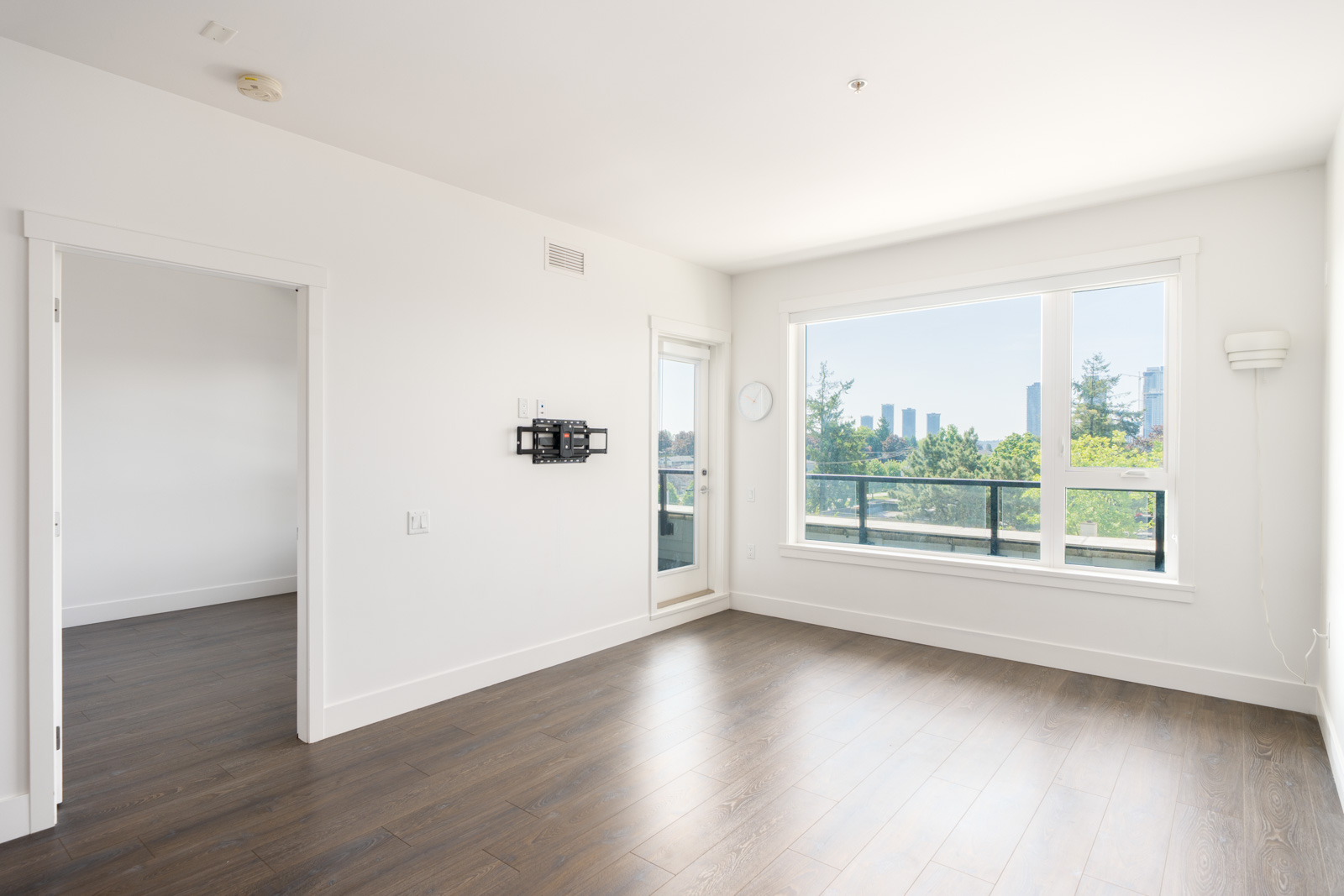 Empty, bright room with white walls, large window, balcony door, and dark wood flooring; doorway leads to an adjacent room. Cityscape visible outside.