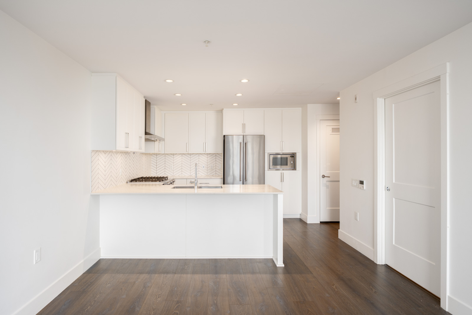 Modern, unfurnished kitchen with white cabinets, stainless steel appliances, a large island, and dark wood flooring in a bright, open space.
