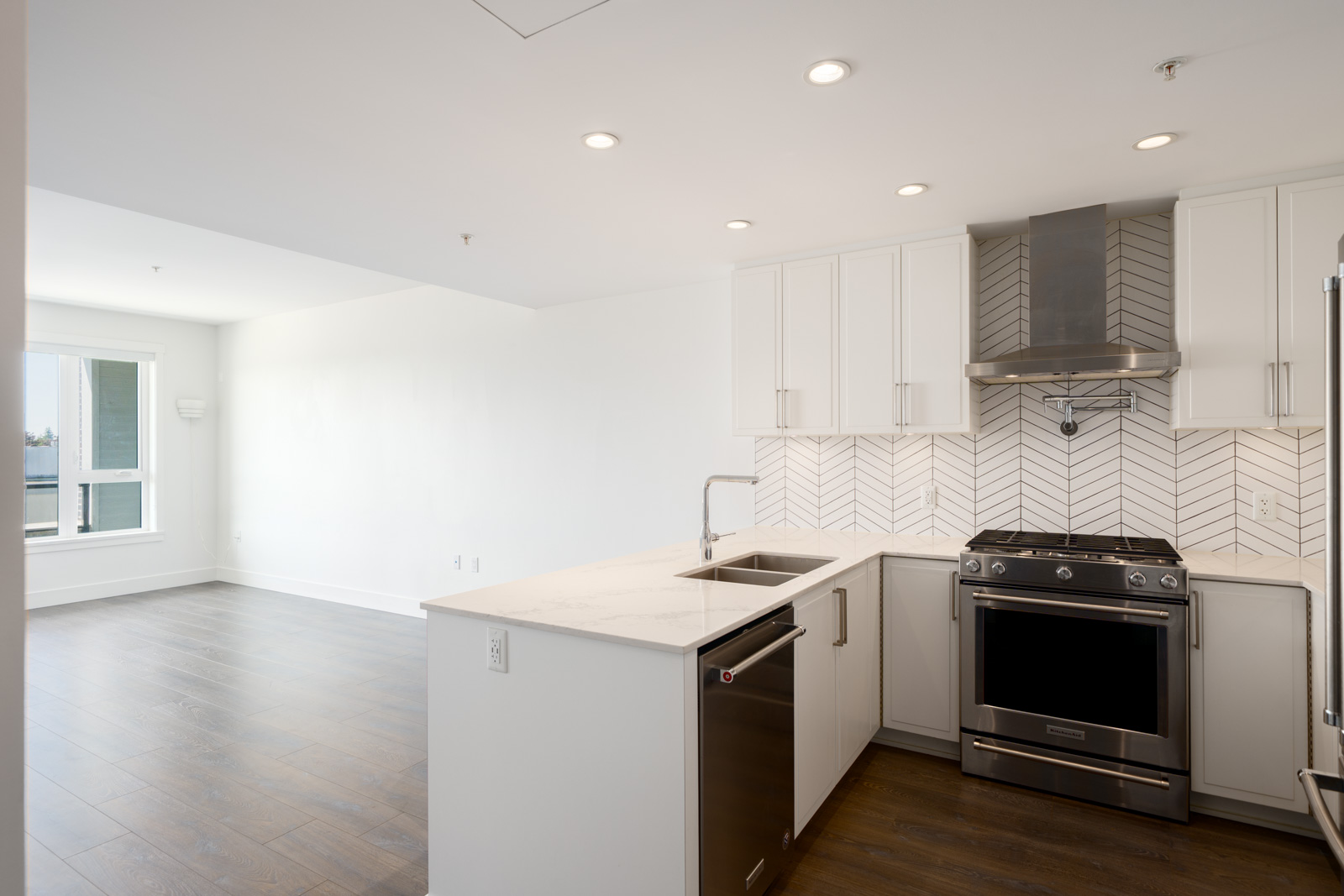 Modern kitchen with white cabinets, stainless steel appliances, a marble countertop, and herringbone tile backsplash, opening to a bright living area with wooden floors.