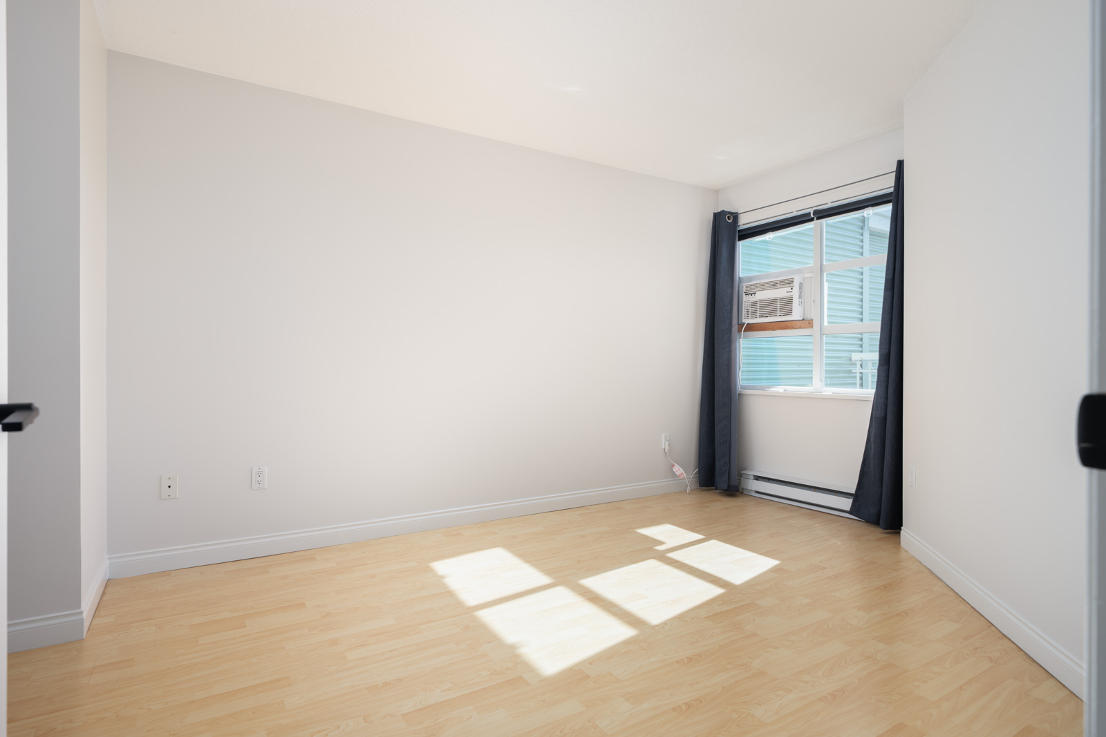 Empty room with light wood flooring, white walls, a window with dark curtains, and a window air conditioning unit. Sunlight creates a bright patch on the floor.