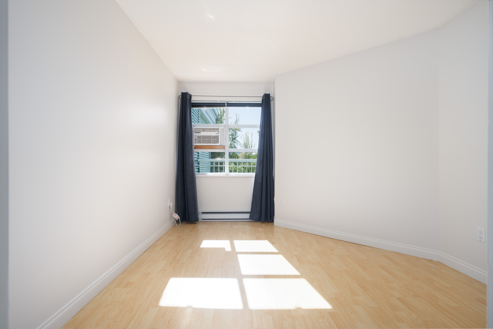 Empty room with light wood flooring, white walls, a window with dark curtains, and sunlight streaming in, casting shadows on the floor.