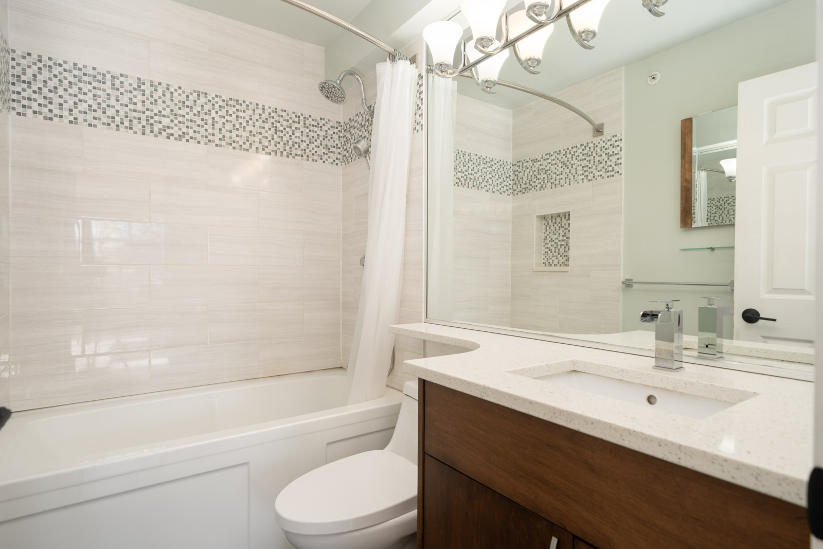Modern bathroom with a bathtub and shower, white tile walls with mosaic accents, a large mirror, countertop sink, and wooden vanity.