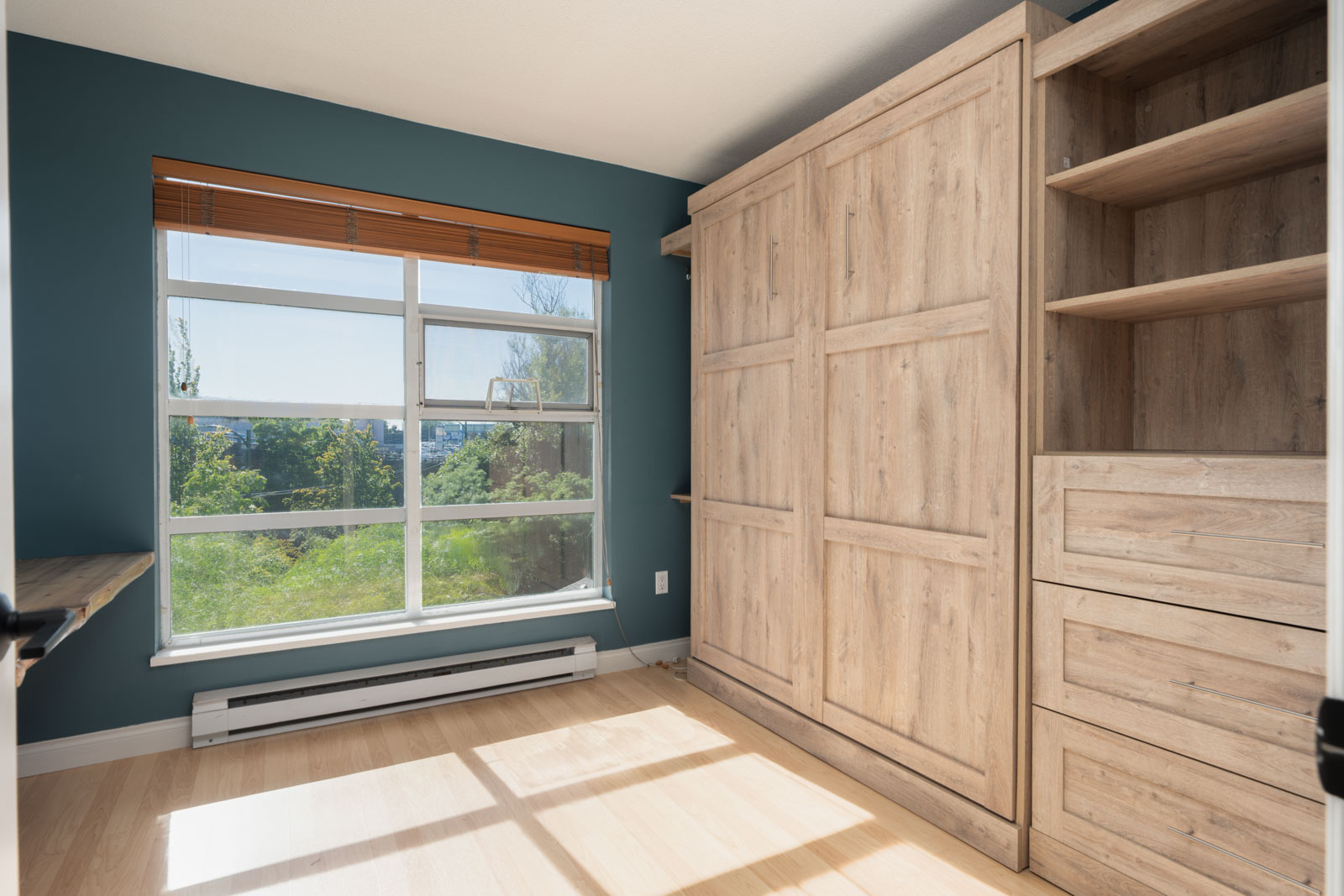A bright room with teal walls, a large window with wooden blinds, and built-in wood cabinets and shelves along one wall.