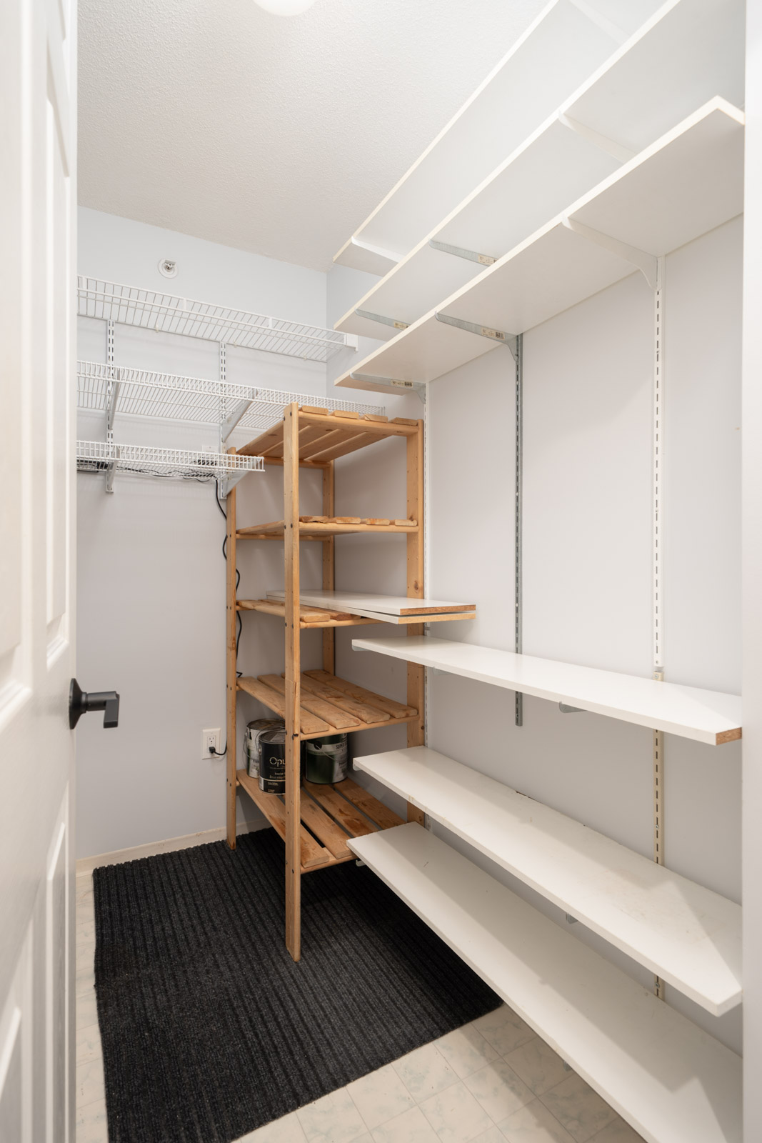 A small walk-in closet with white shelves, a wooden shelving unit, wire racks, and a black floor mat.