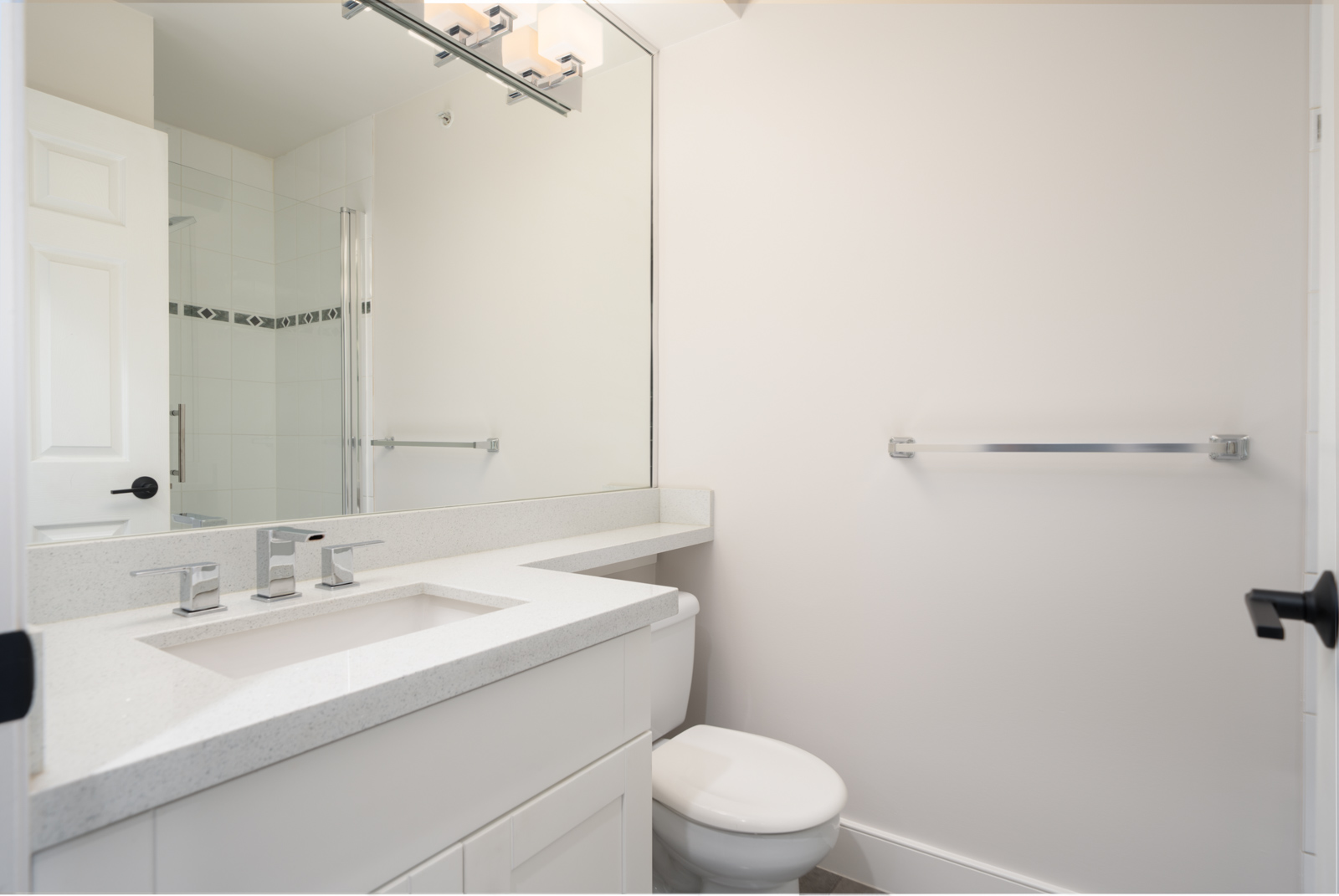 Modern bathroom with white walls, a large mirror, a sink with a rectangular countertop, a toilet, glass shower, and a towel bar on the wall.
