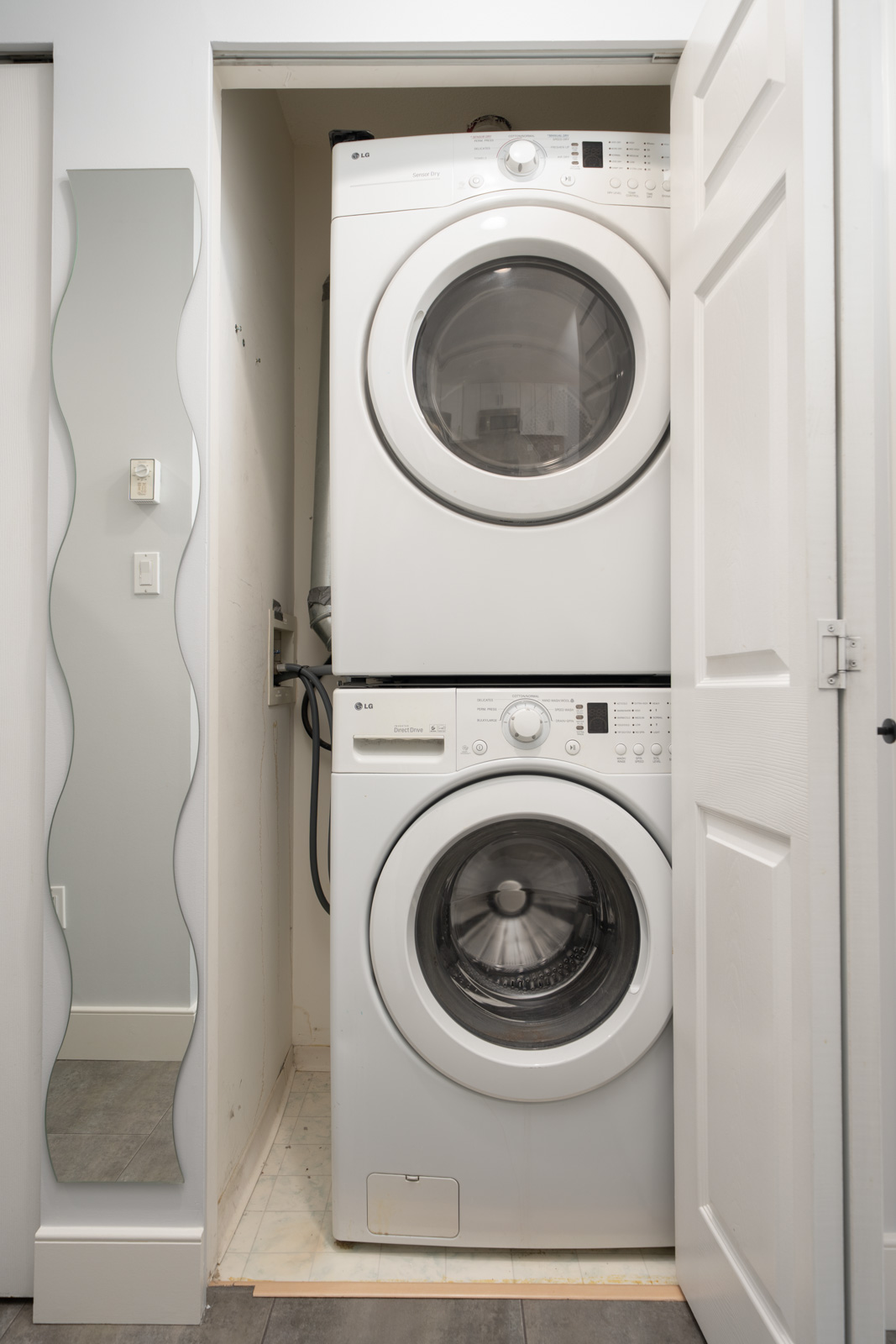 Stacked front-load washer and dryer in a small laundry closet with white walls and a wavy full-length mirror on the left.