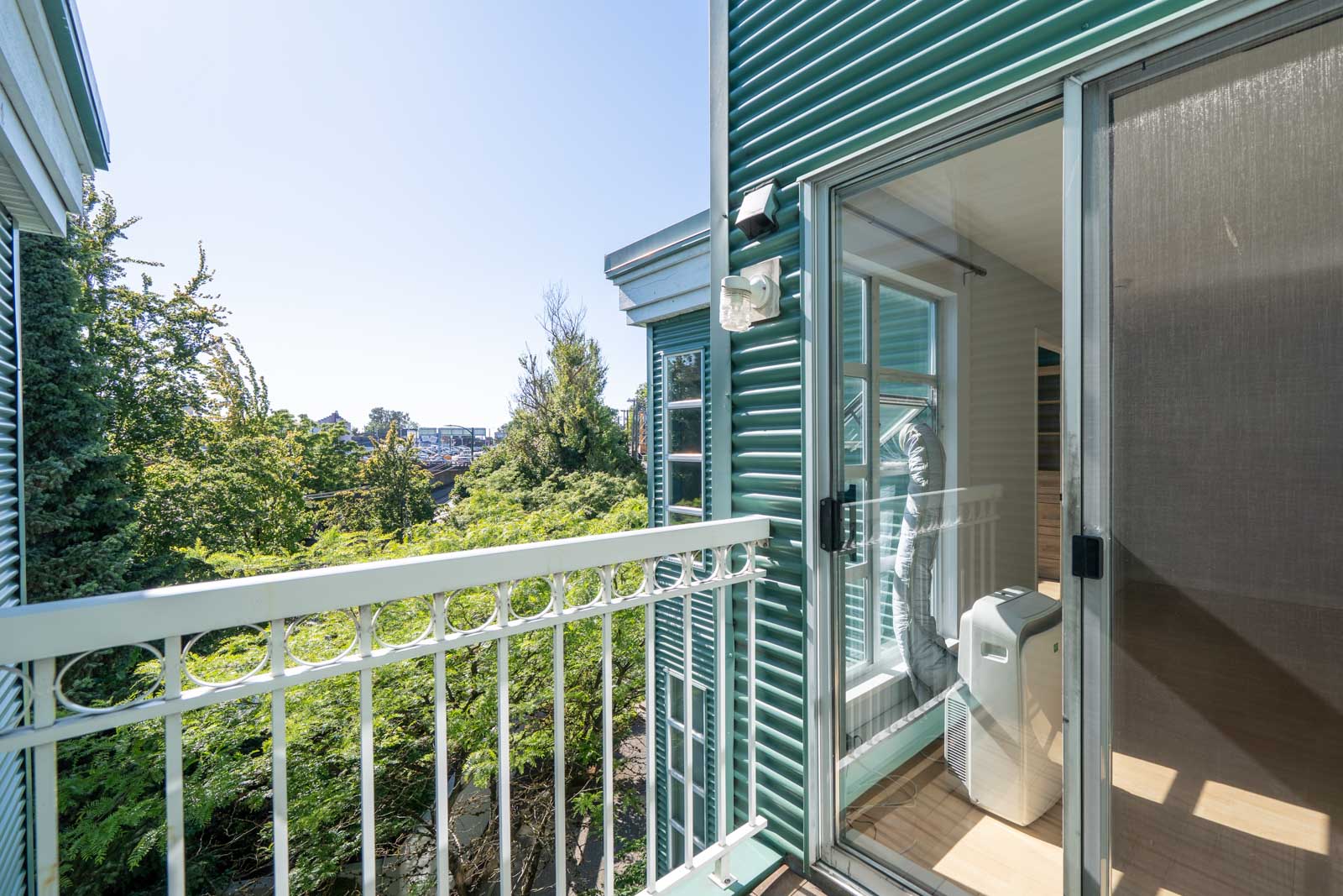 A small balcony with a white railing and green siding, connected to a room with a portable air conditioner by a sliding glass door; greenery and blue sky in the background.