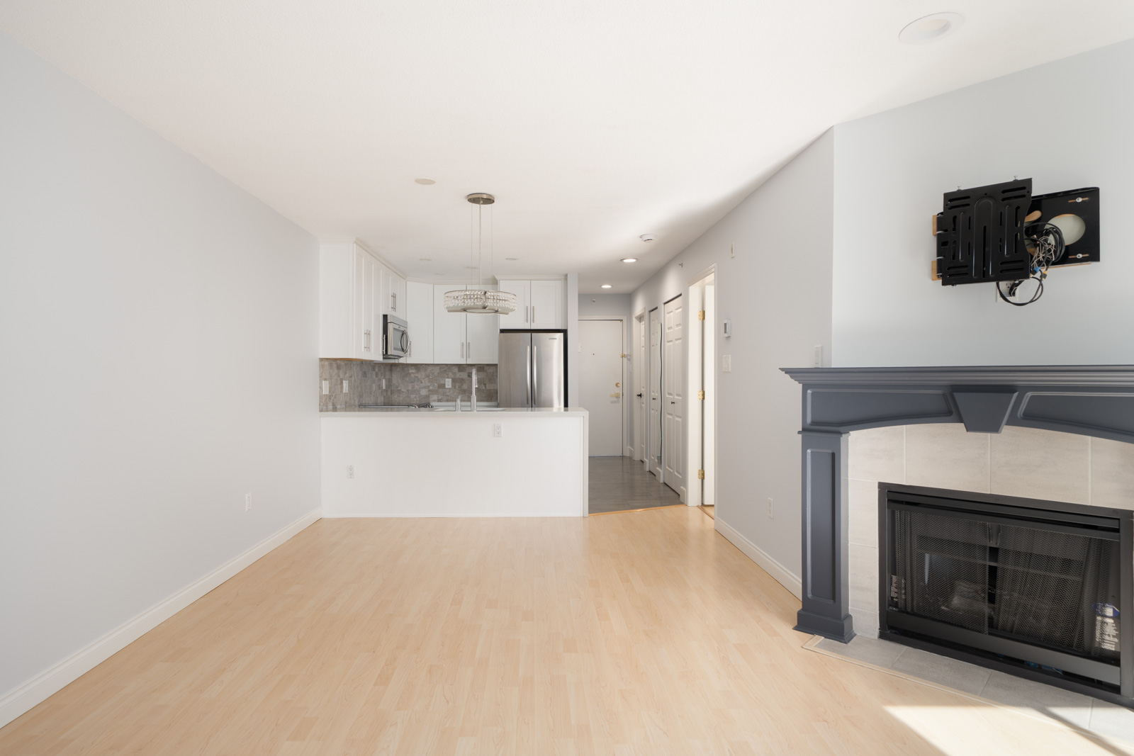 Bright, empty living area with light wood flooring, a gray fireplace, wall-mounted TV bracket, and a view into a modern white kitchen with stainless steel appliances.