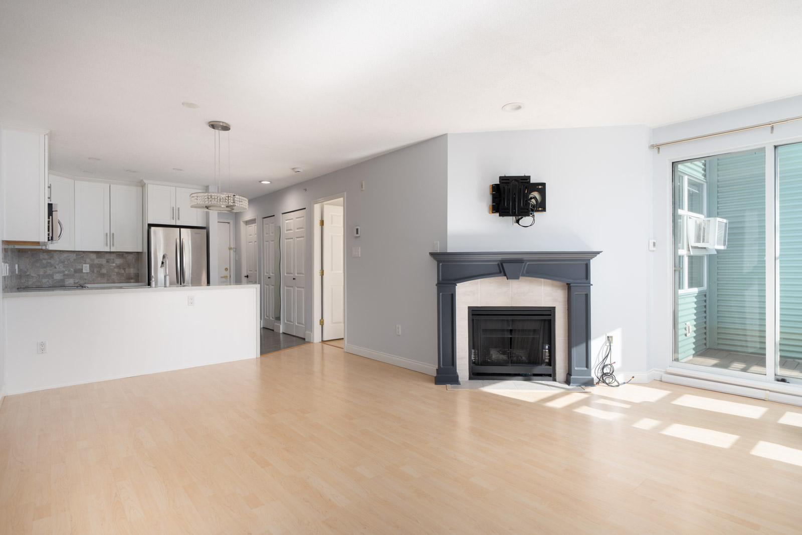 Bright, unfurnished living room with light wood floors, a black fireplace, and sliding glass doors; adjacent kitchen features white cabinets and stainless steel appliances.