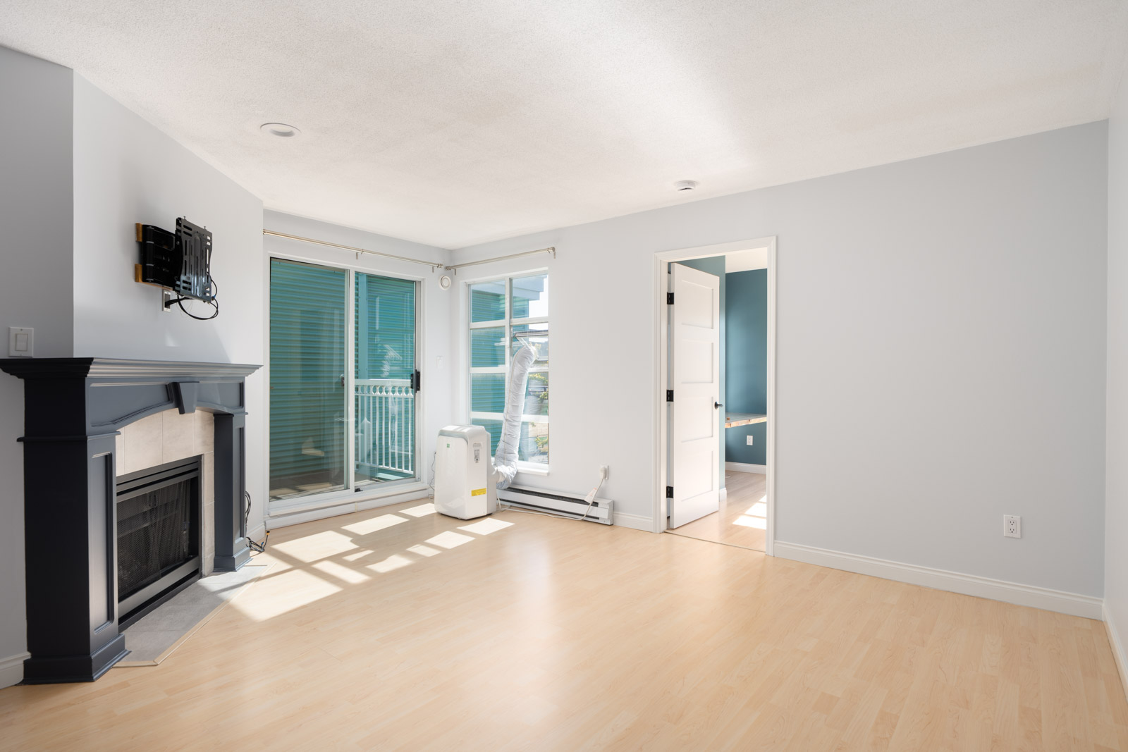 Empty living room with light wood floors, a black fireplace, wall-mounted TV, large windows, and an open doorway leading to another room.