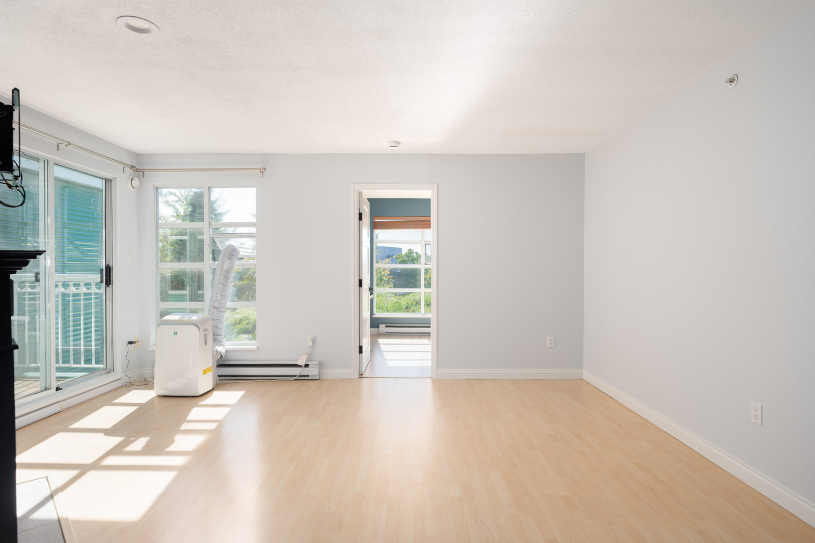 Bright, empty living room with light wood flooring, white walls, large windows, a glass door to a balcony, and an open doorway leading to another sunlit room.