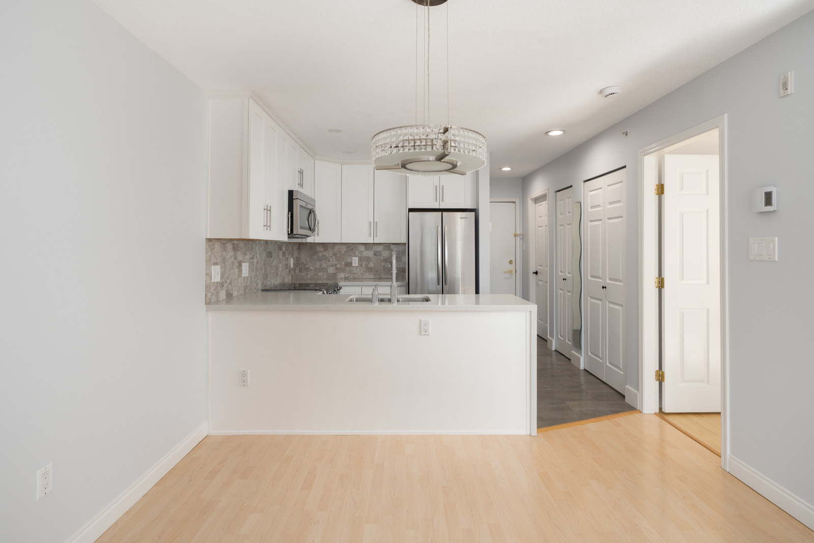 Modern kitchen with white cabinets, stainless steel appliances, light wood flooring, tile backsplash, and a pendant light fixture above a white countertop. Hallway with multiple doors visible.